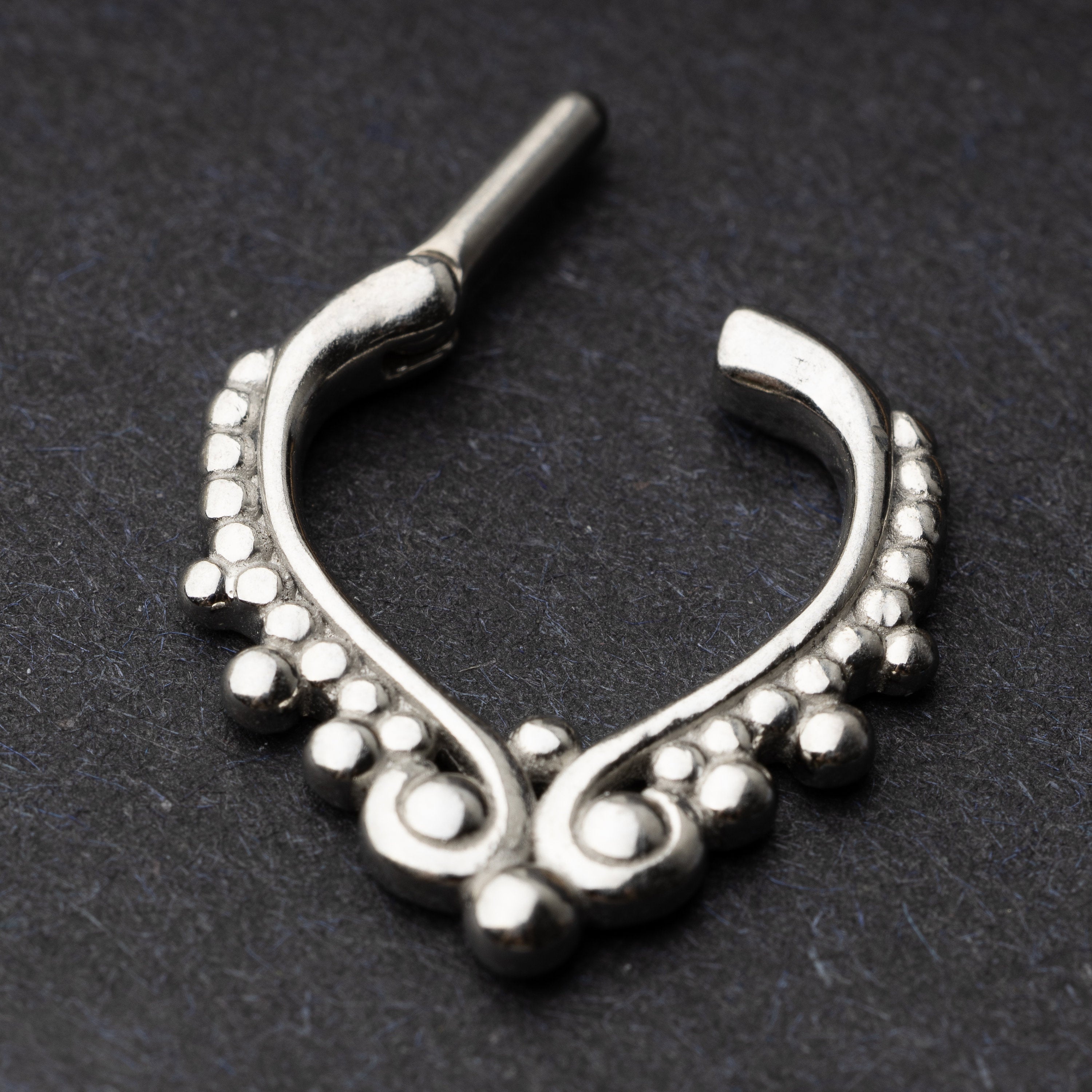 Close-up of a silver septum clicker with a teardrop shape and beaded embellishments, displayed on a dark background with a secure hinged clasp which is open in this image.