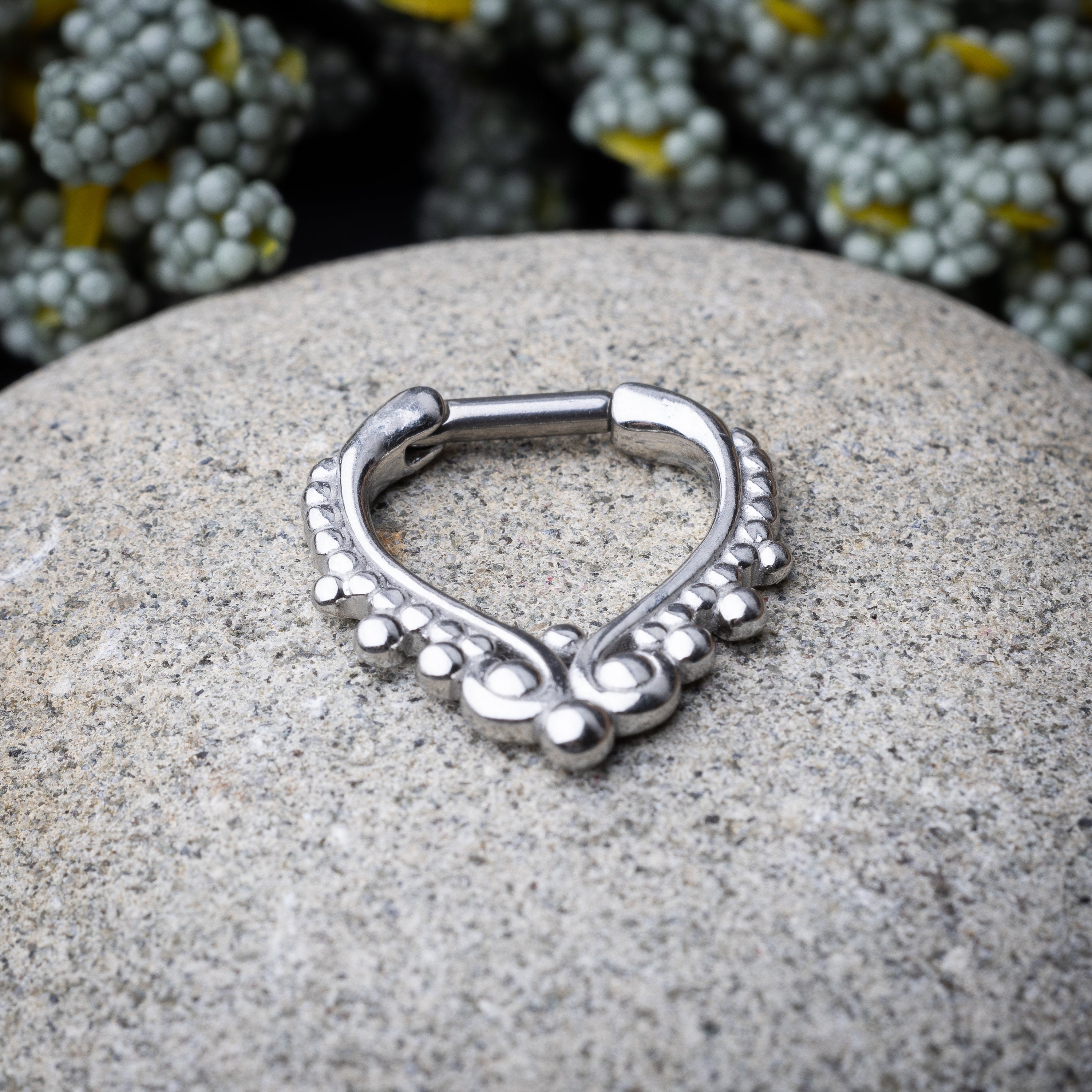 Close-up of a silver septum clicker with a teardrop shape and beaded embellishments, displayed on a stone background with a secure hinged clasp.