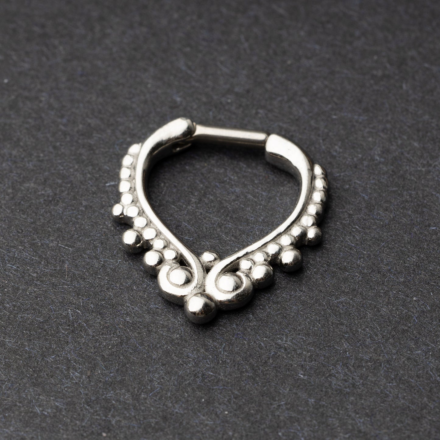 Close-up of a silver septum clicker with a teardrop shape and beaded embellishments, displayed on a dark background with a secure hinged clasp.