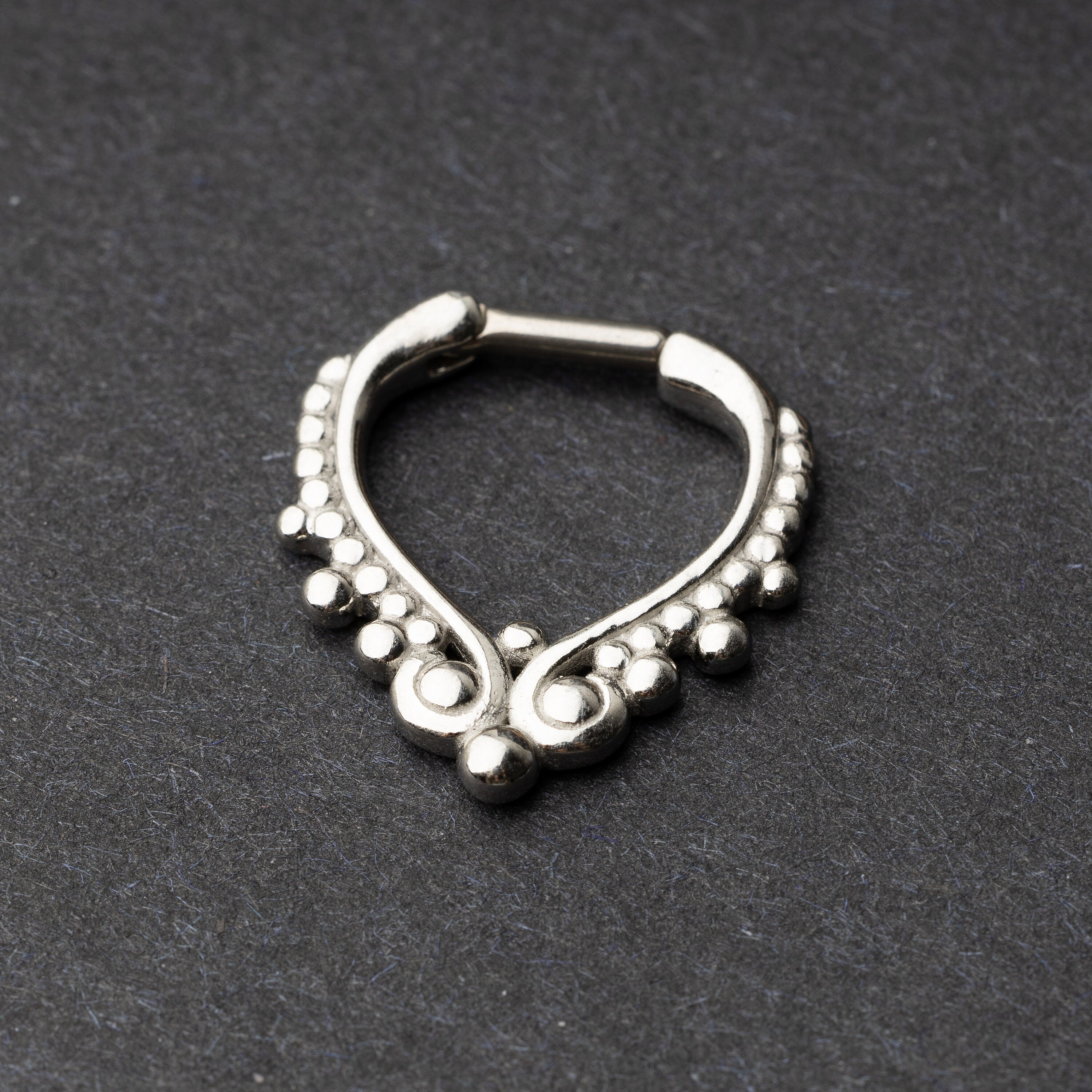 Close-up of a silver septum clicker with a teardrop shape and beaded embellishments, displayed on a dark background with a secure hinged clasp.