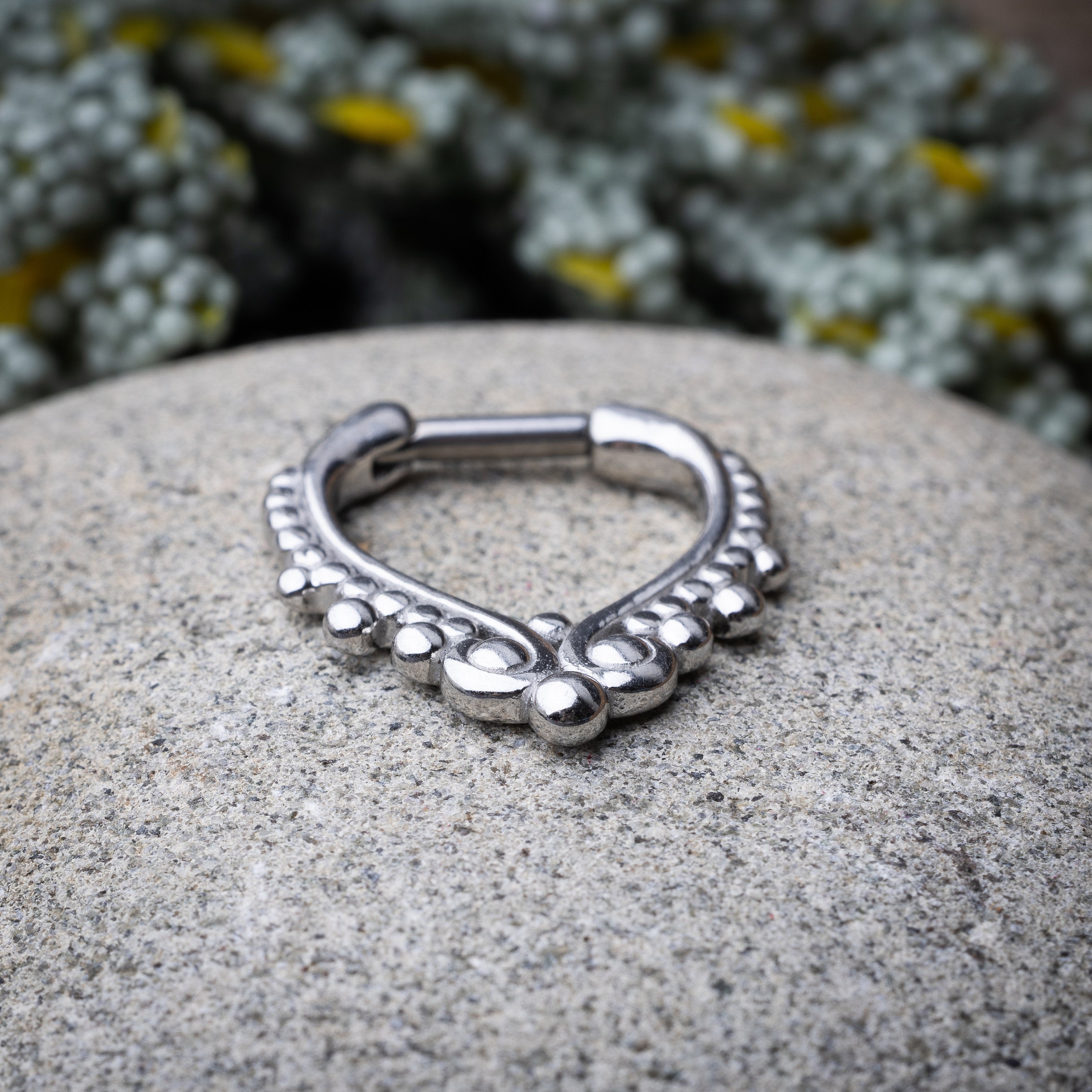 Close-up of a silver septum clicker with a teardrop shape and beaded embellishments, displayed on a stone background with a secure hinged clasp.