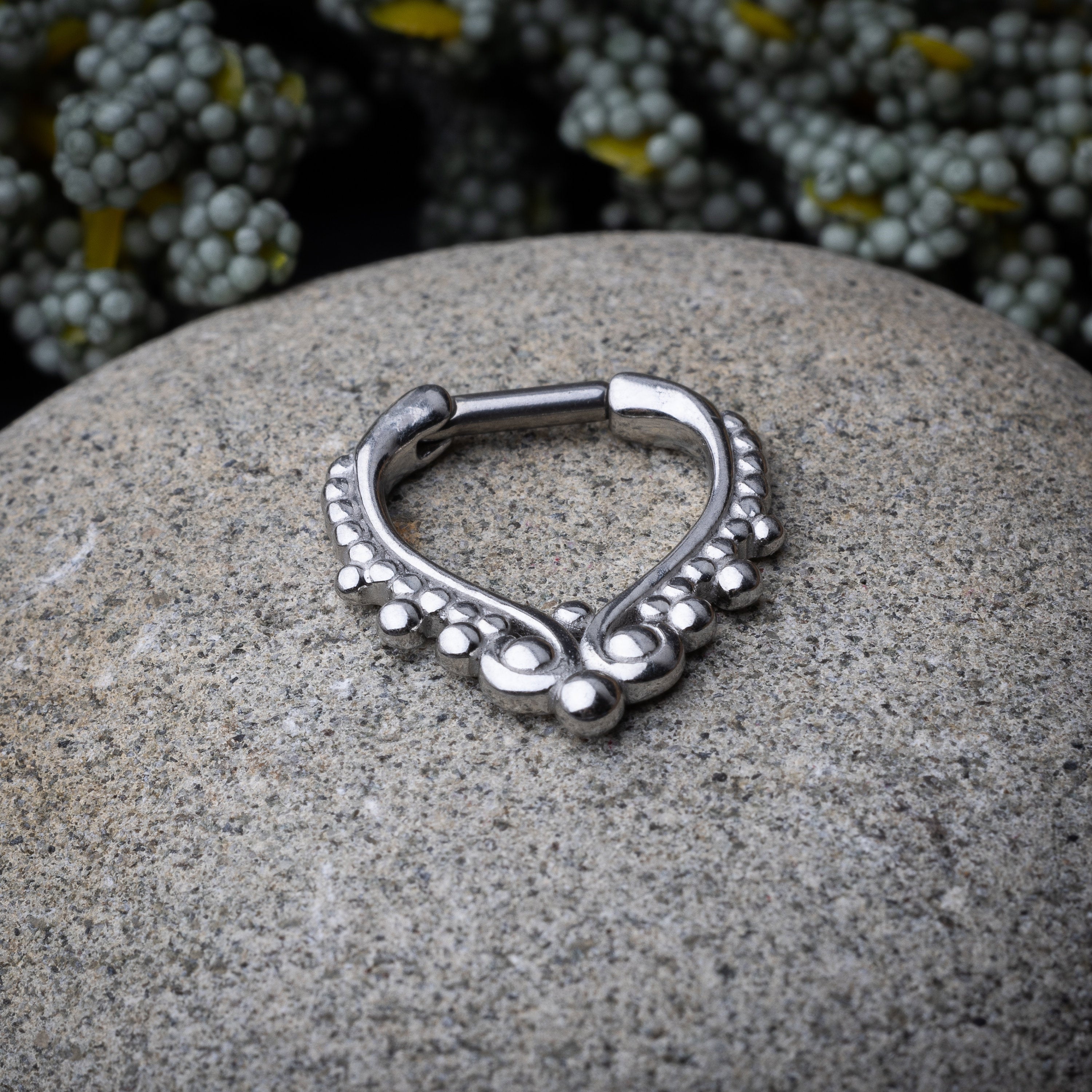 Close-up of a silver septum clicker with a teardrop shape and beaded embellishments, displayed on a stone background with a secure hinged clasp.