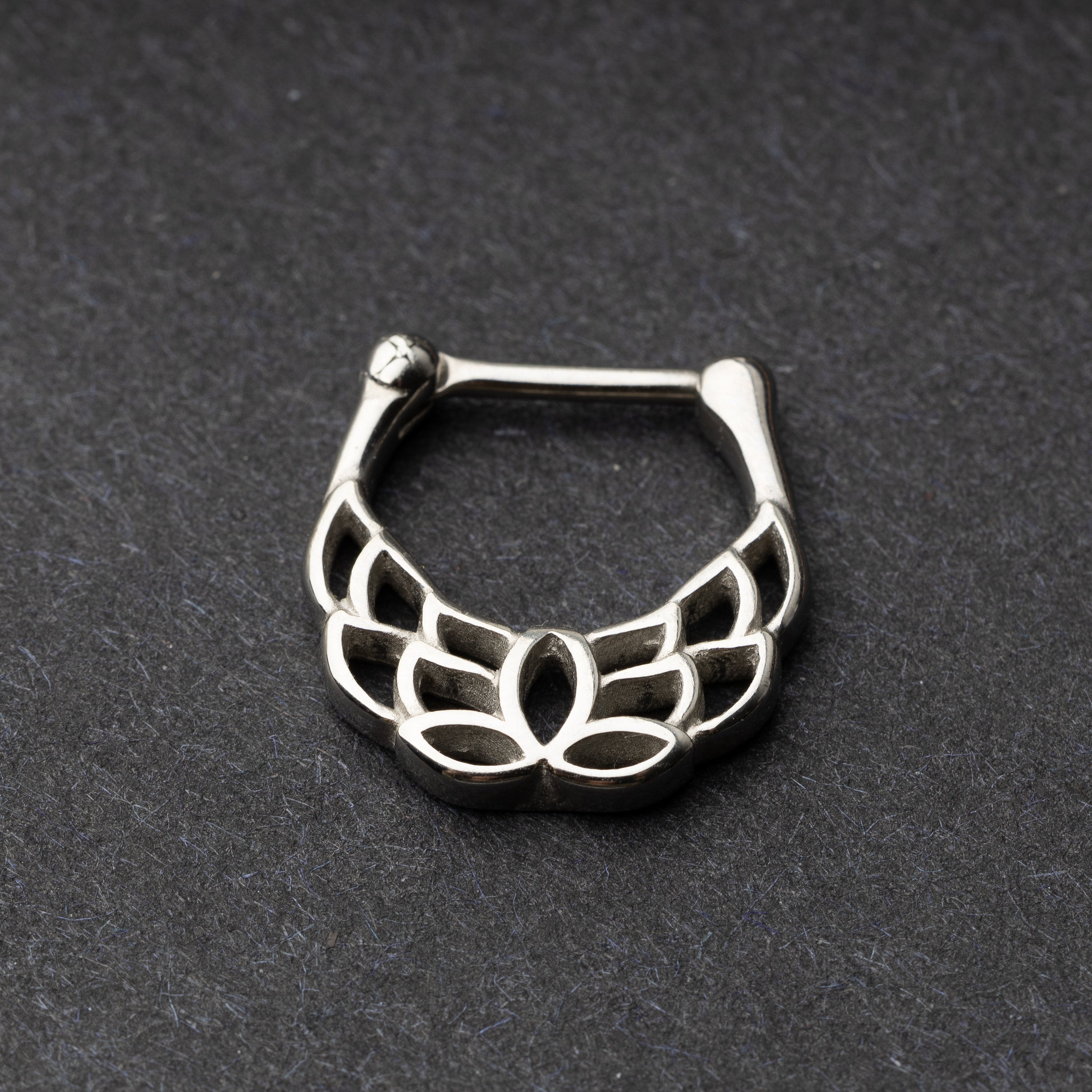 Close-up of a silver septum clicker with a lotus-inspired design, showcasing intricate petal details and a secure hinged clasp on a dark background.