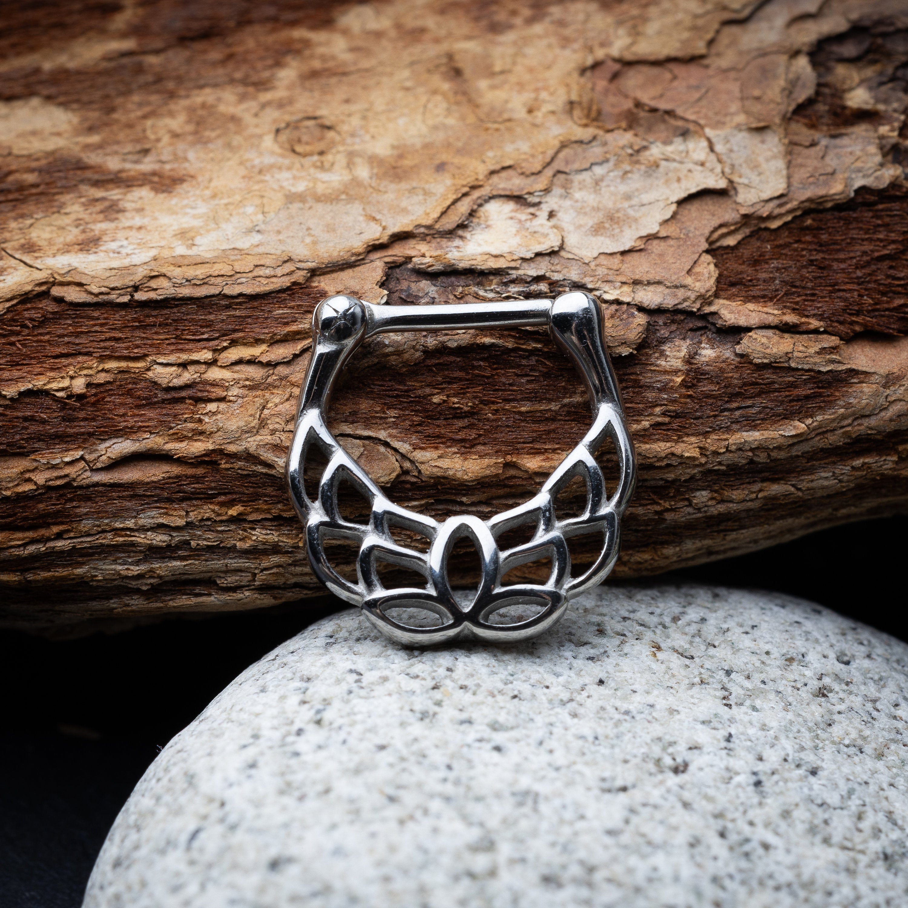 Close-up of a silver septum clicker with a lotus-inspired design, showcasing intricate petal details and a secure hinged clasp on a stone and wooden background.