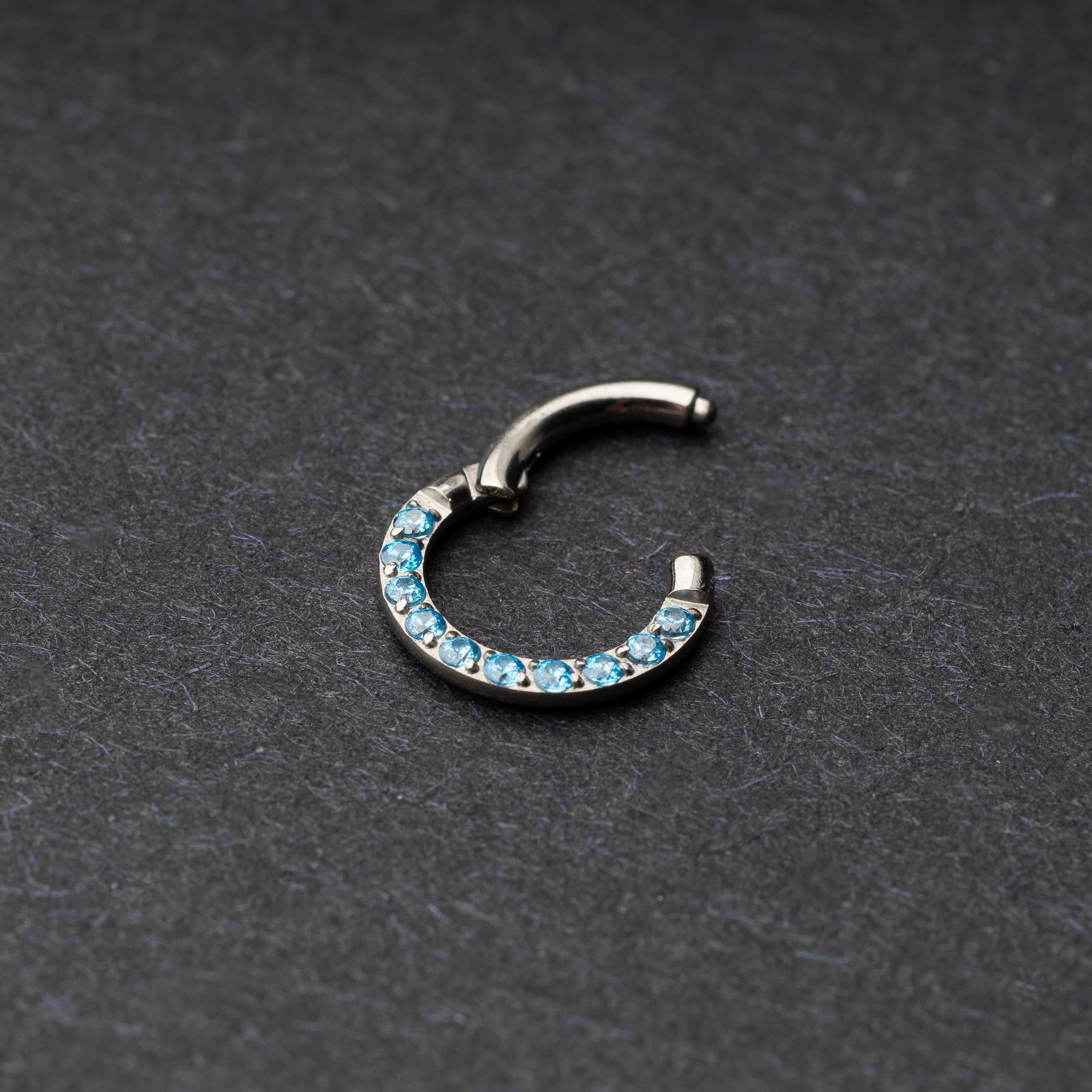 Stainless steel septum clicker with bright blue gemstones, displayed on a dark textured background.