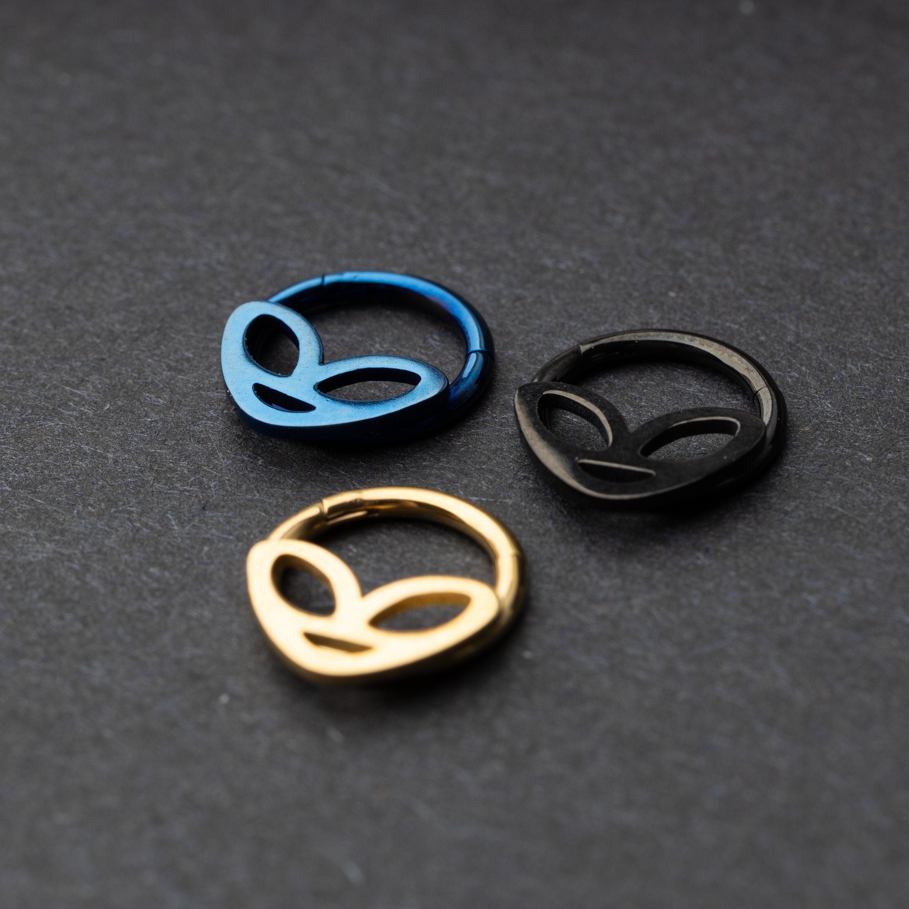 Three alien-themed septum clickers in metallic blue, matte black, and polished gold finishes, displayed on a dark background.