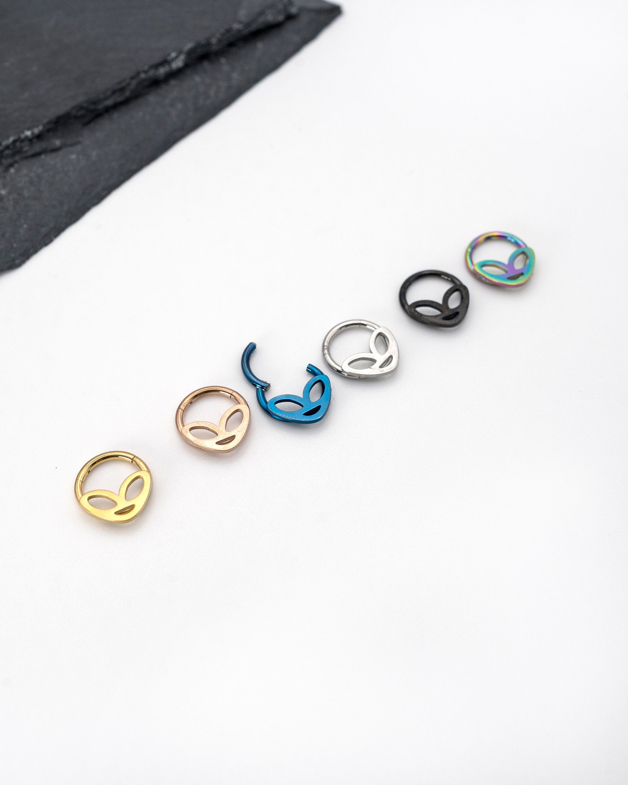 Six alien-themed septum clickers in metallic blue, matte black, silver, rainbow, rose gold and polished gold finishes, displayed on a dark background.