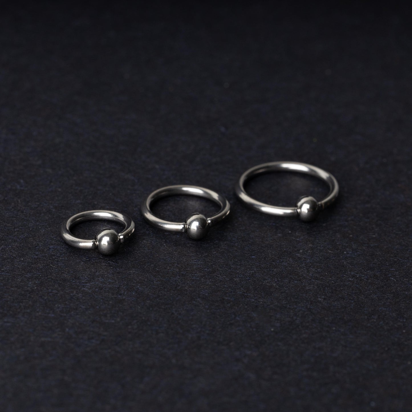 Three polished stainless steel captive bead rings of varying sizes displayed on a dark matte background.