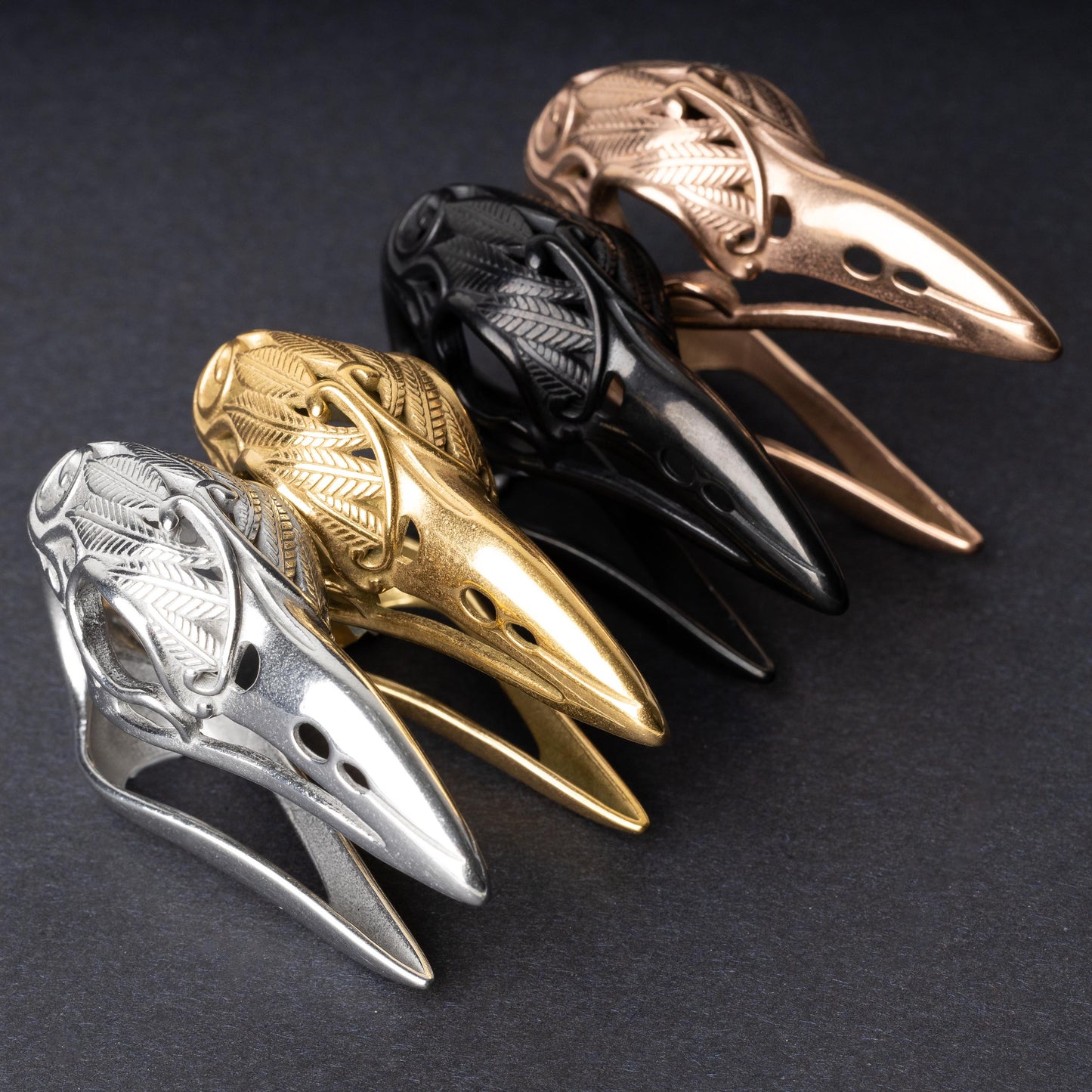 Raven skull ear hangers in silver, gold, black, and rose gold finishes, showcasing intricate feather engravings and polished details, displayed on a dark background.