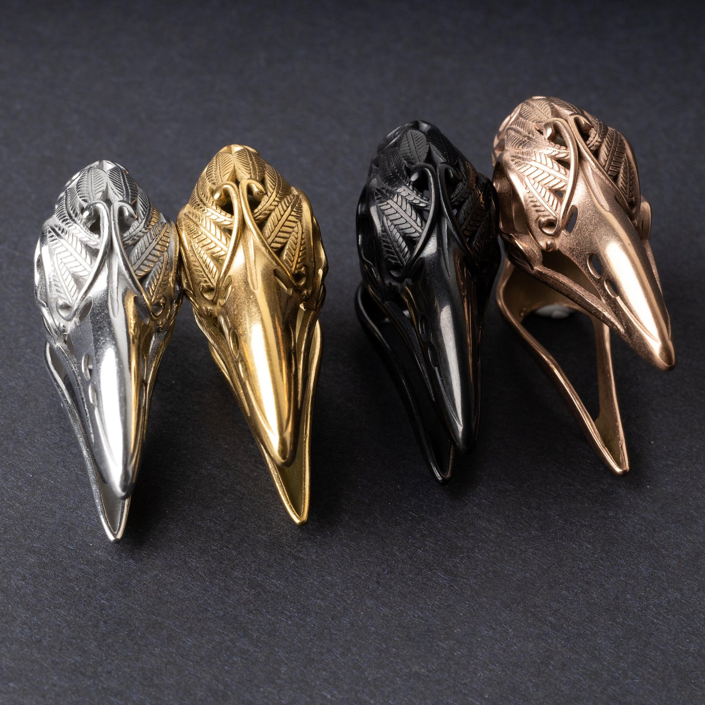 raven skull ear hangers in silver, gold, black, and rose gold finishes, showcasing intricate feather engravings and polished details, displayed on a dark background.