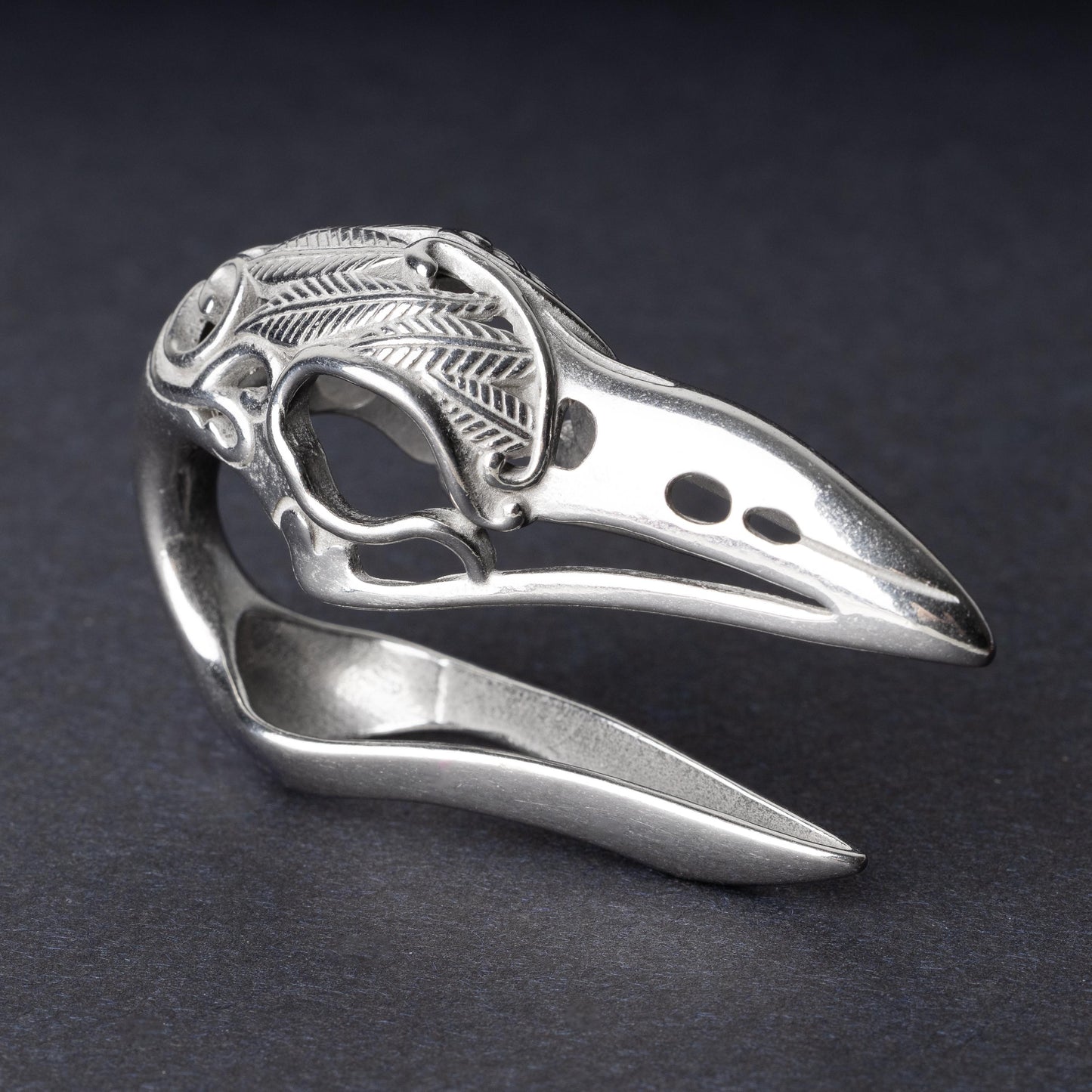 A silver raven skull ear hanger showcasing intricate feather engravings and polished details, displayed on a dark background.