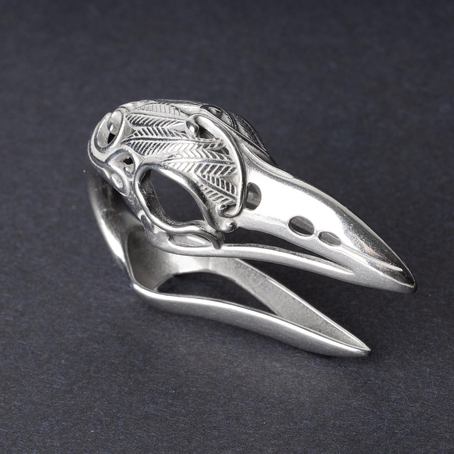 A silver raven skull ear hanger showcasing intricate feather engravings and polished details, displayed on a dark background.