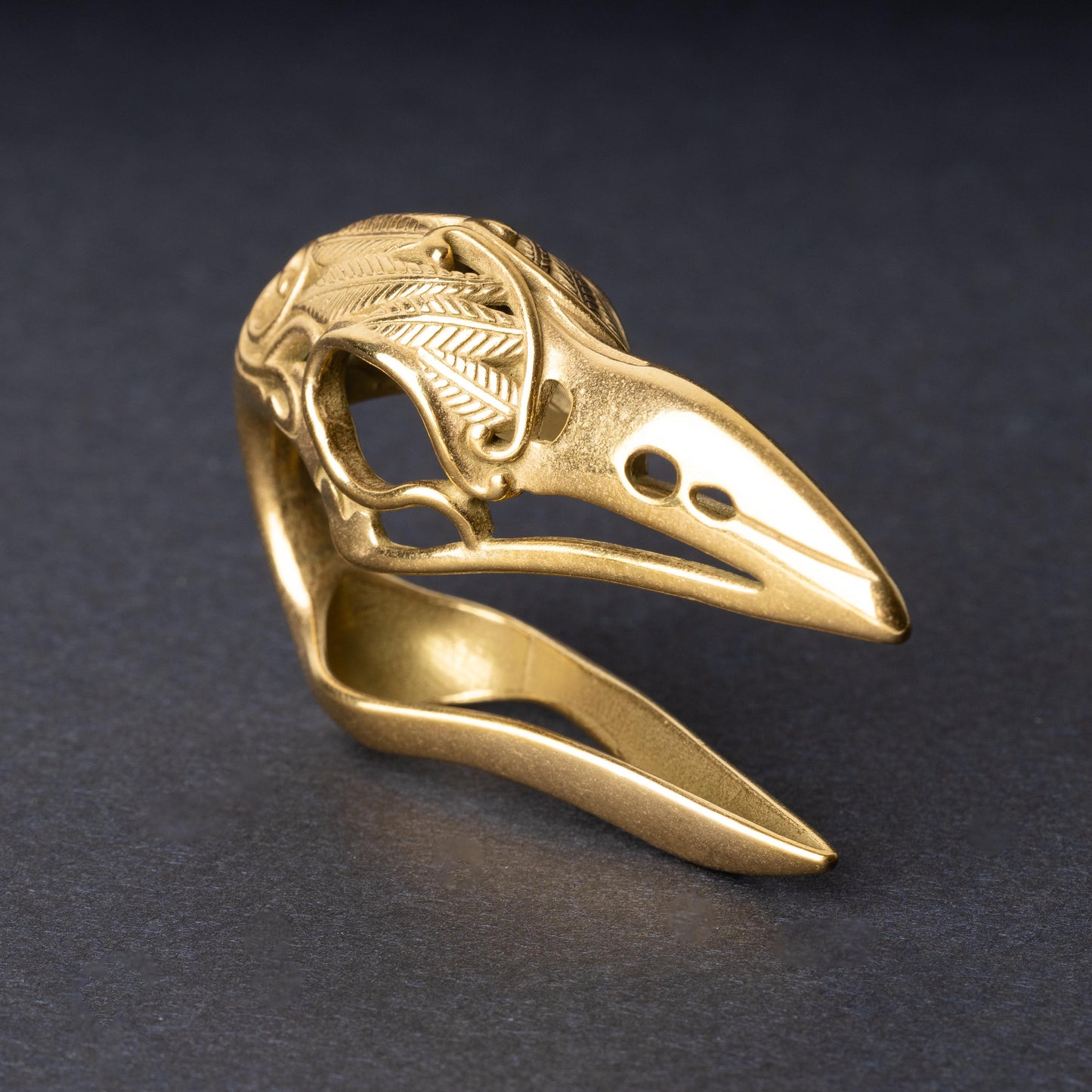 A gold raven skull ear hanger showcasing intricate feather engravings and polished details, displayed on a dark background.