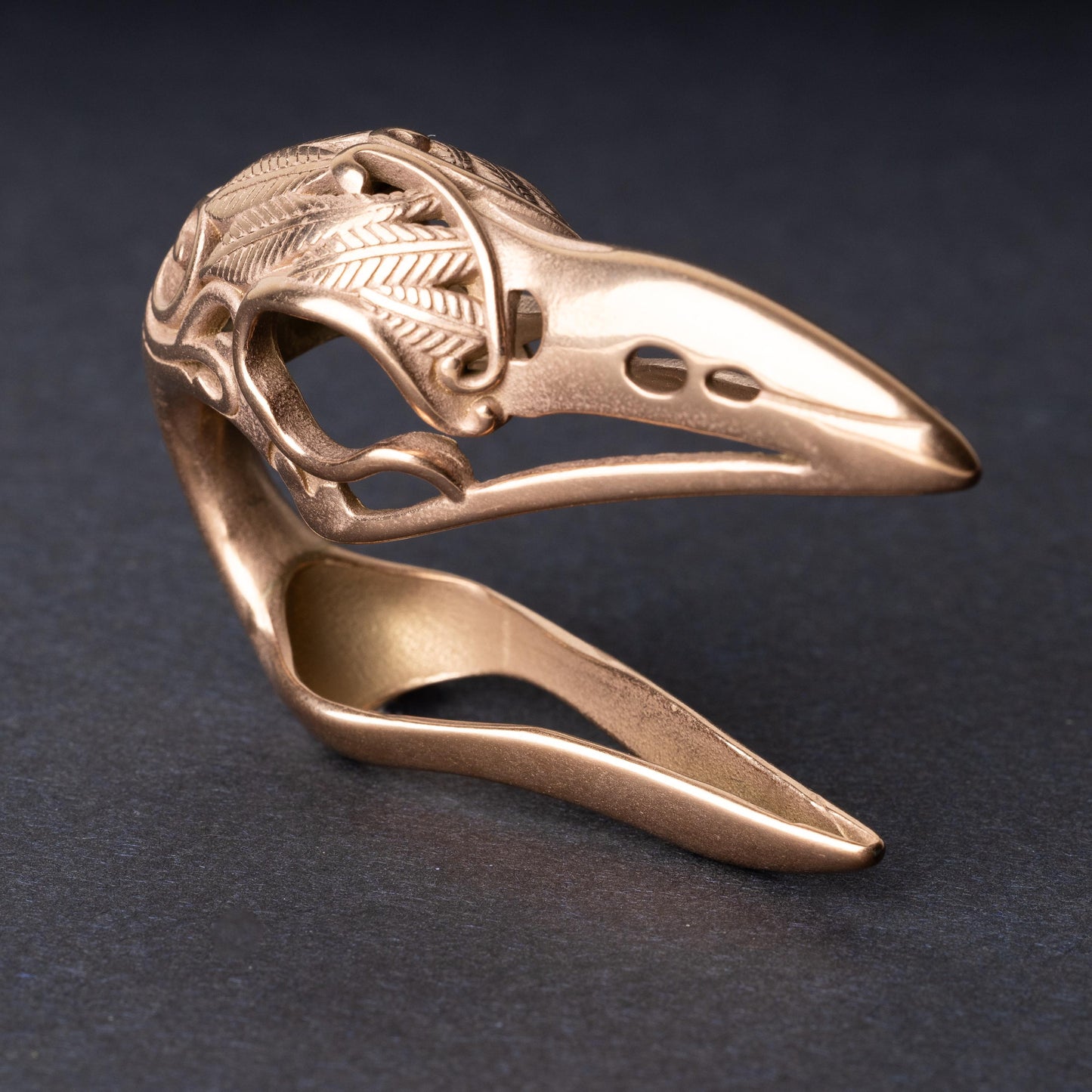 A rose gold raven skull ear hanger showcasing intricate feather engravings and polished details, displayed on a dark background.