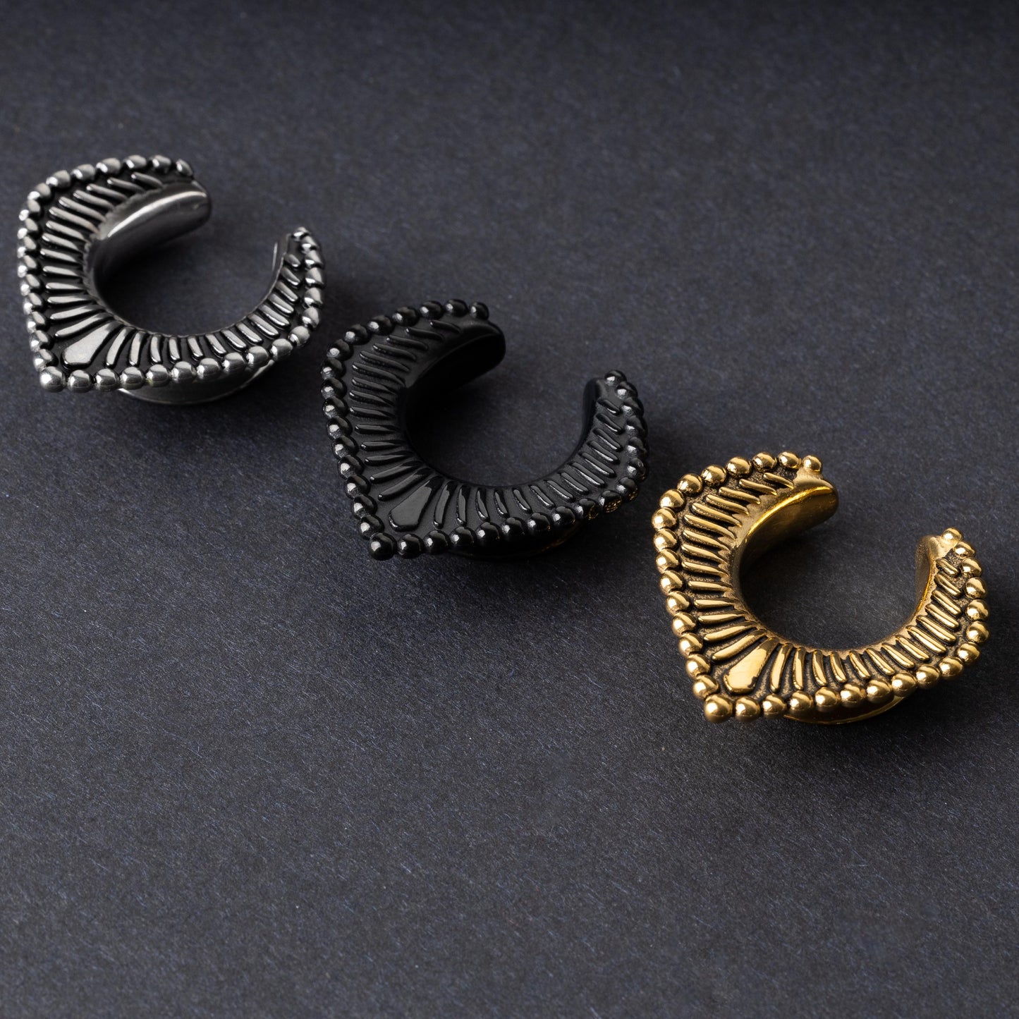 Three V-shaped stainless steel clickers in silver, black, and gold finishes arranged on a dark textured background.