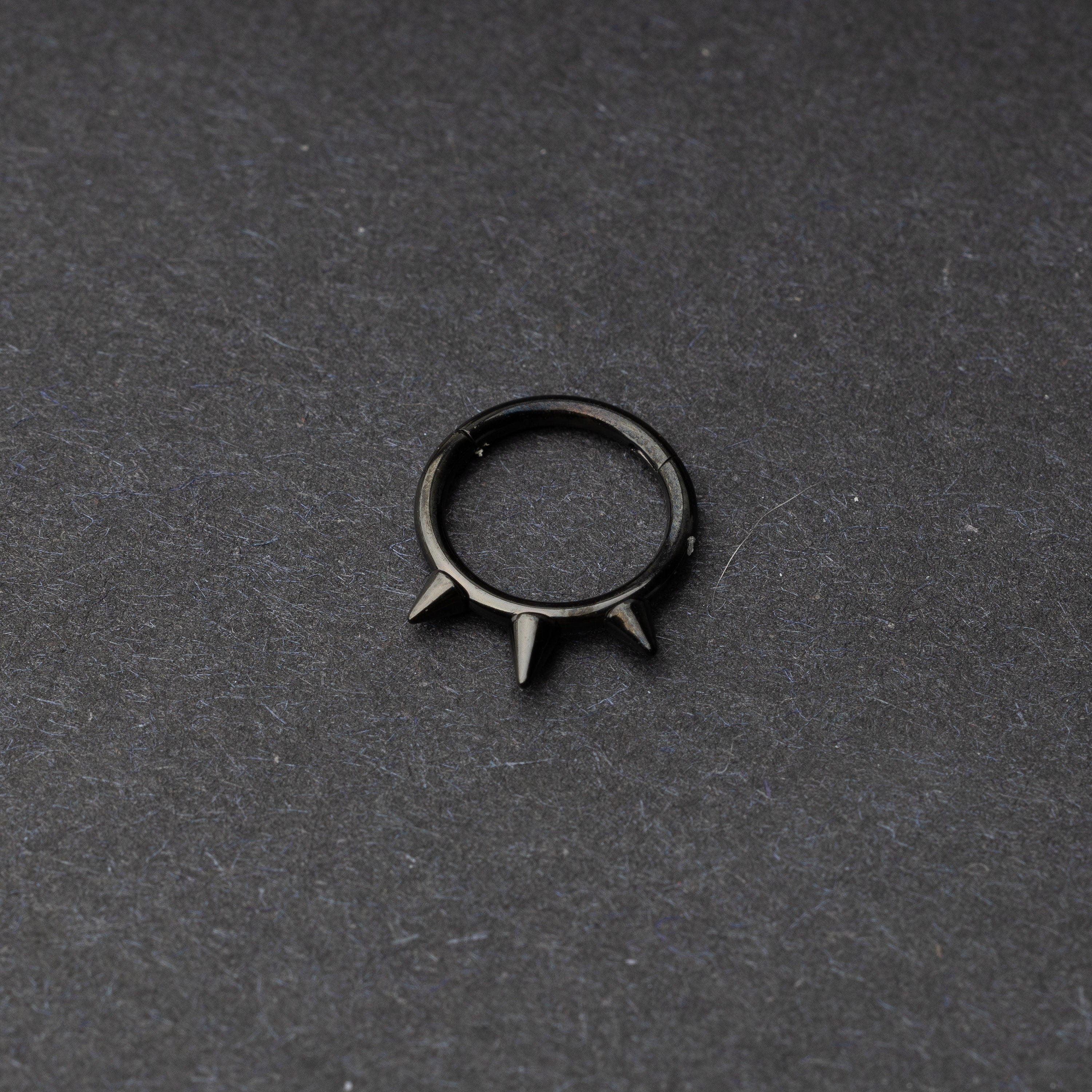 Black-tone stainless steel clicker ring with three spikes, displayed on a dark textured background.