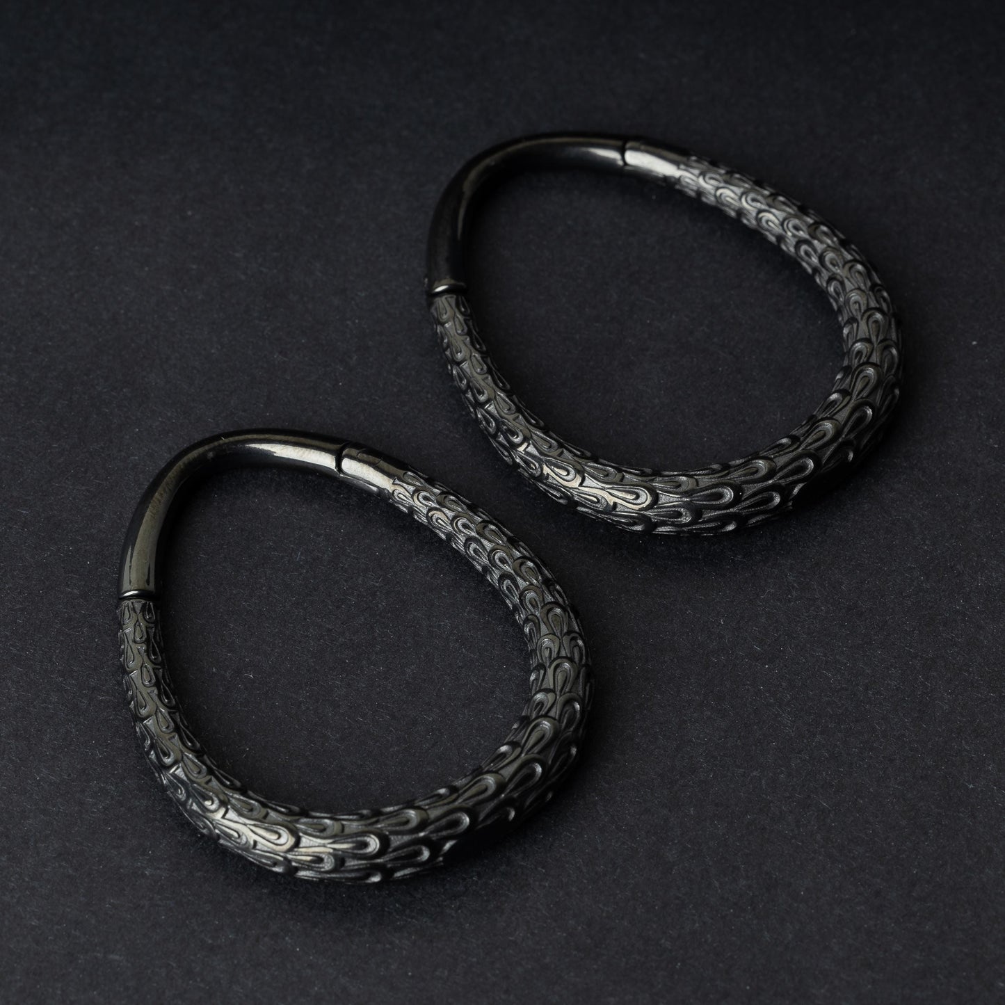A pair of black teardrop ear weights featuring a detailed dragon-scale texture and a seamless hinge closure, displayed on a dark background.