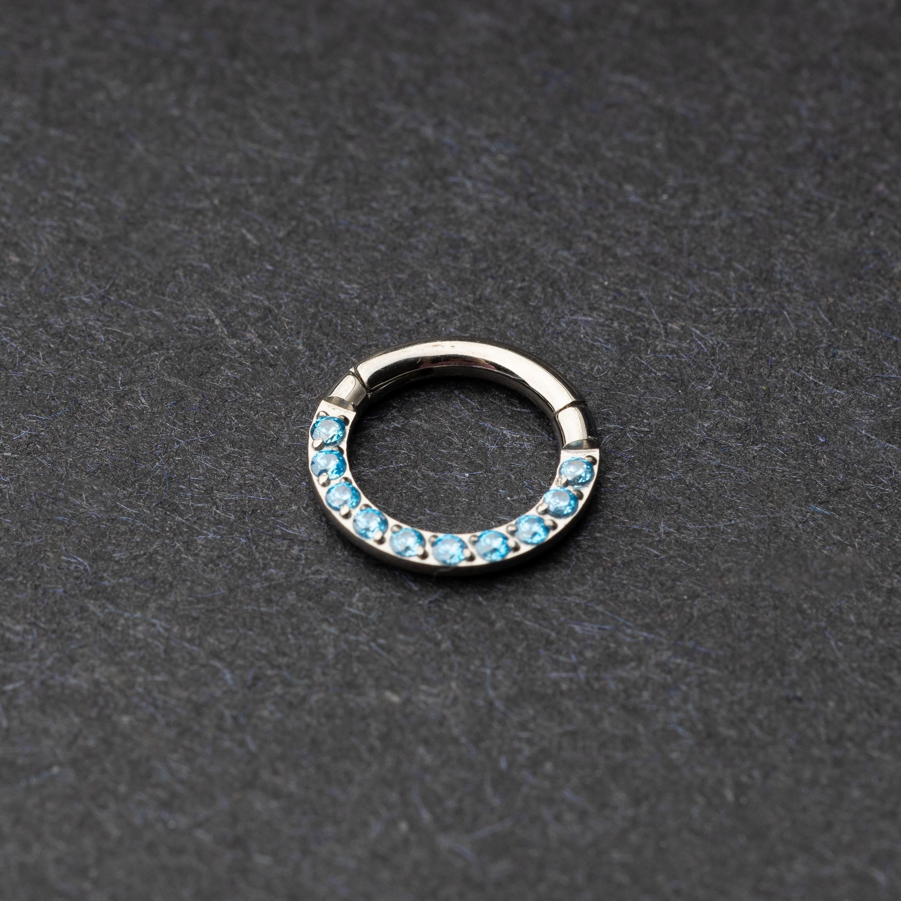 Stainless steel septum clicker with bright blue gemstones, displayed on a dark textured background.