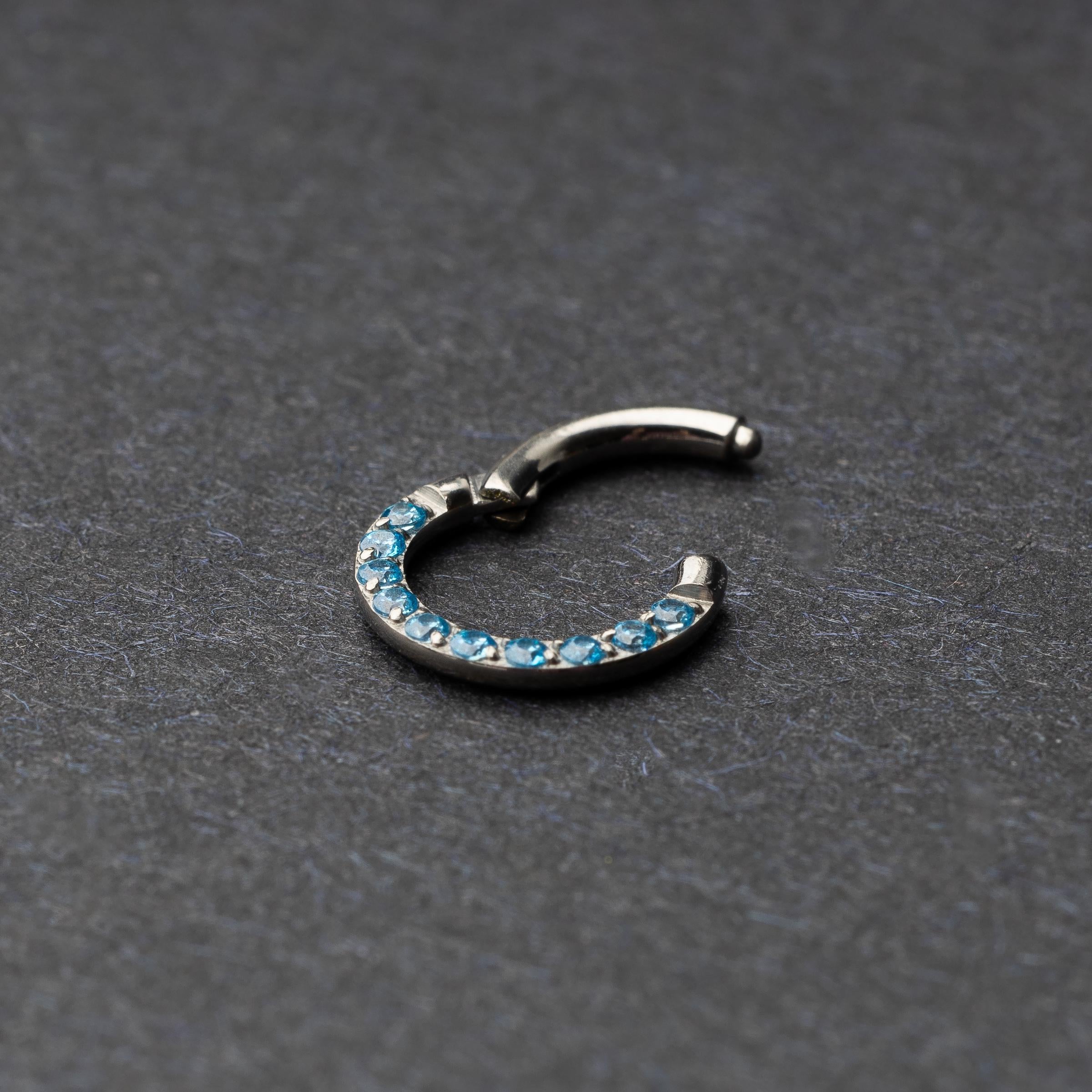 Stainless steel septum clicker with bright blue gemstones, displayed on a dark textured background.