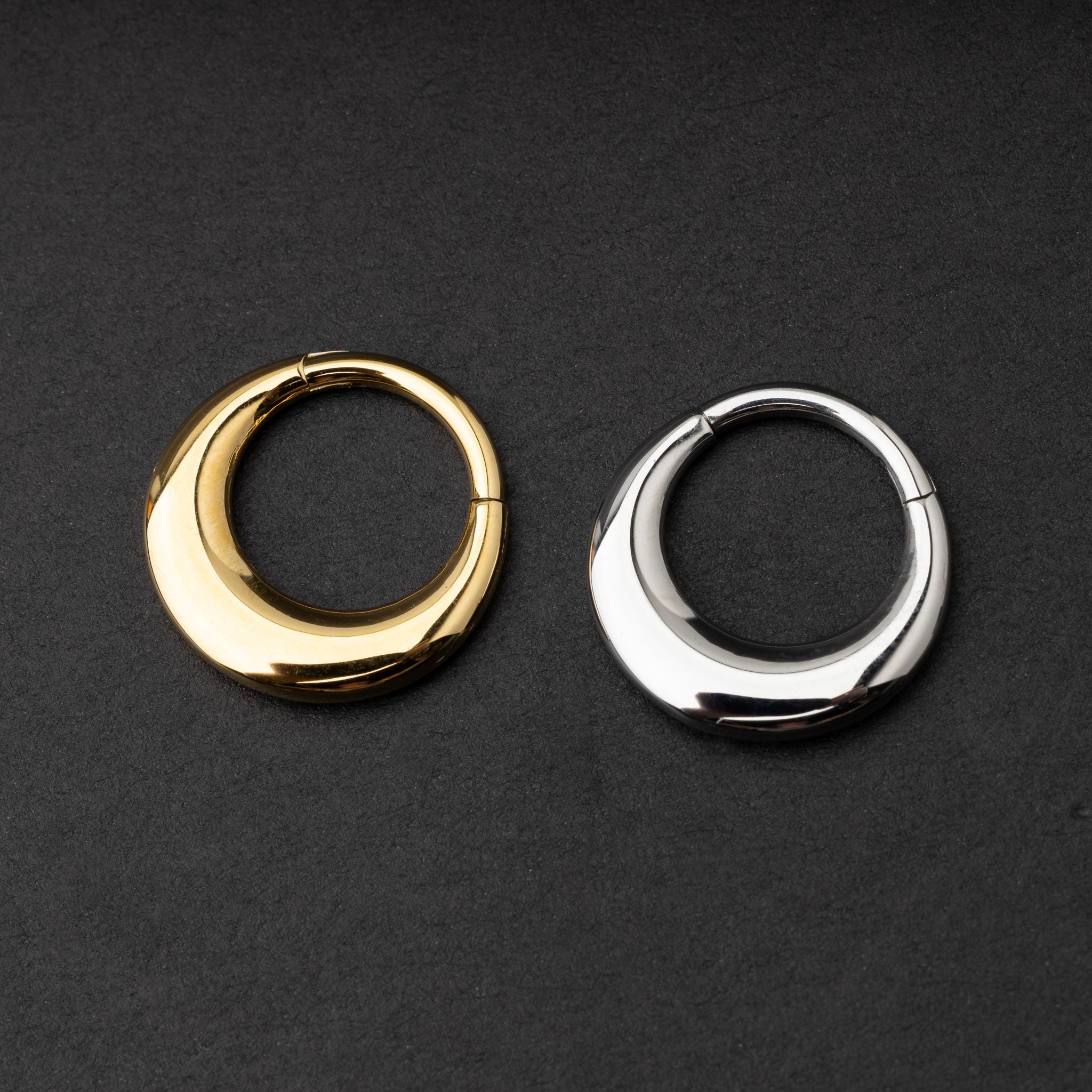Crescent-shaped septum clickers, one in gold and one in silver, with a polished finish, displayed on a dark background.
