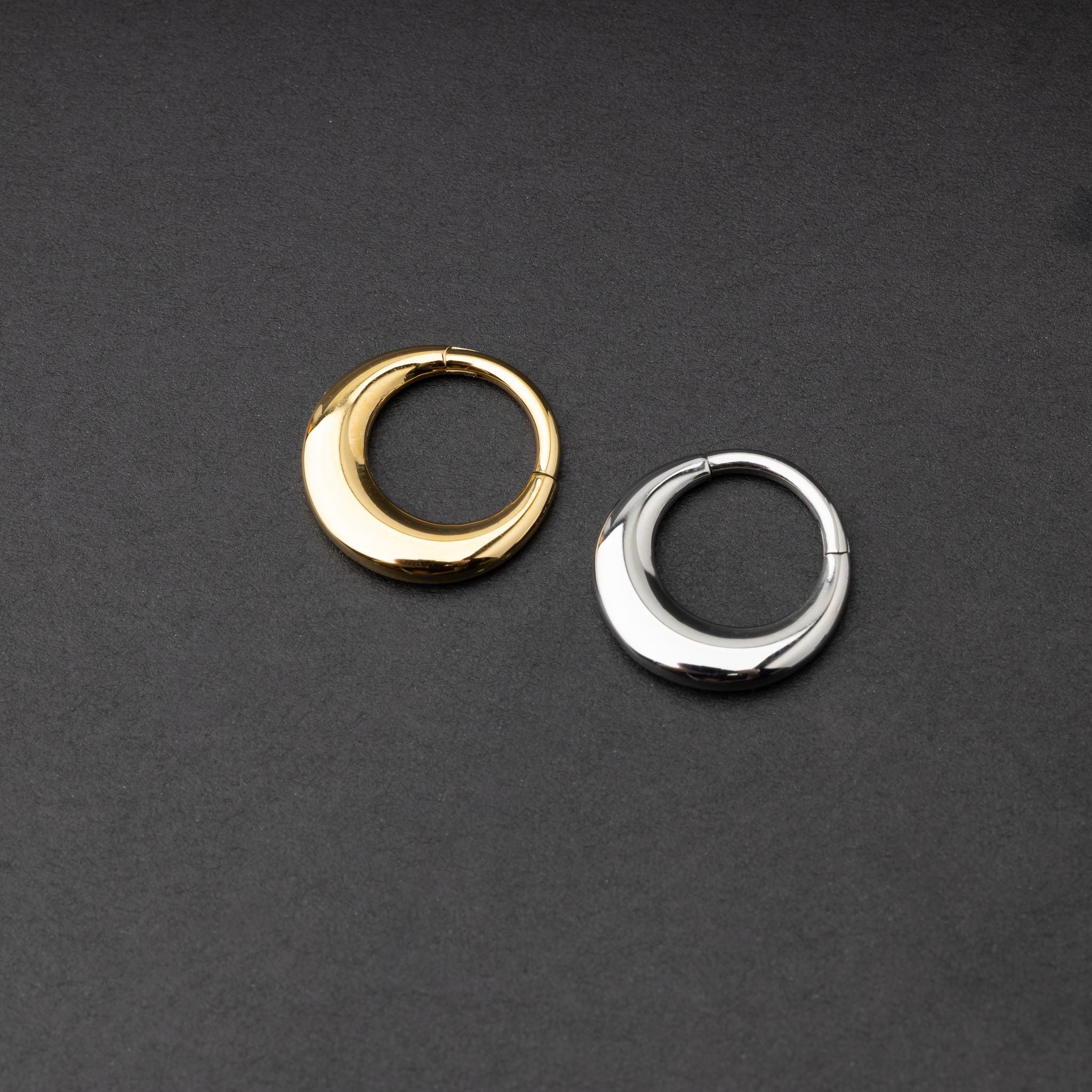 Crescent-shaped septum clickers, one in gold and one in silver, with a polished finish, displayed on a dark background.