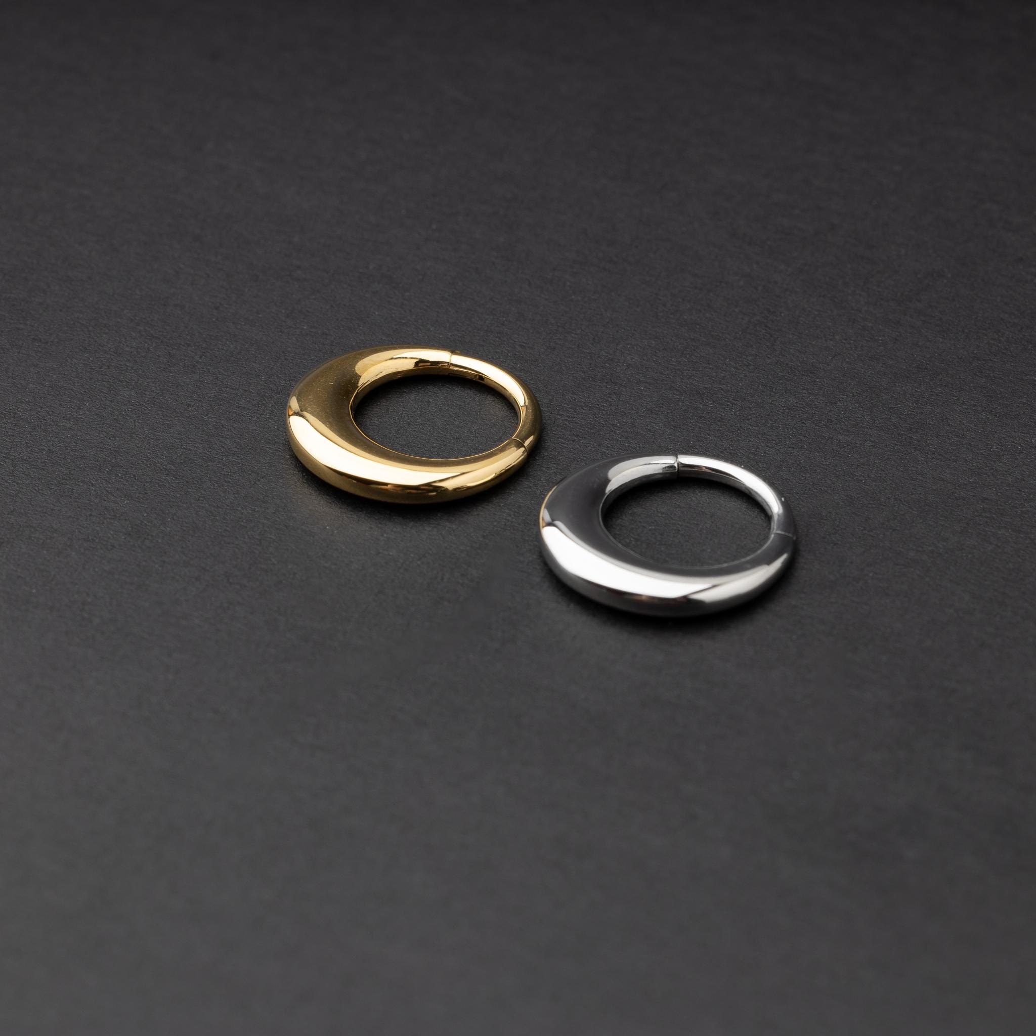 Crescent-shaped septum clickers, one in gold and one in silver, with a polished finish, displayed on a dark background.