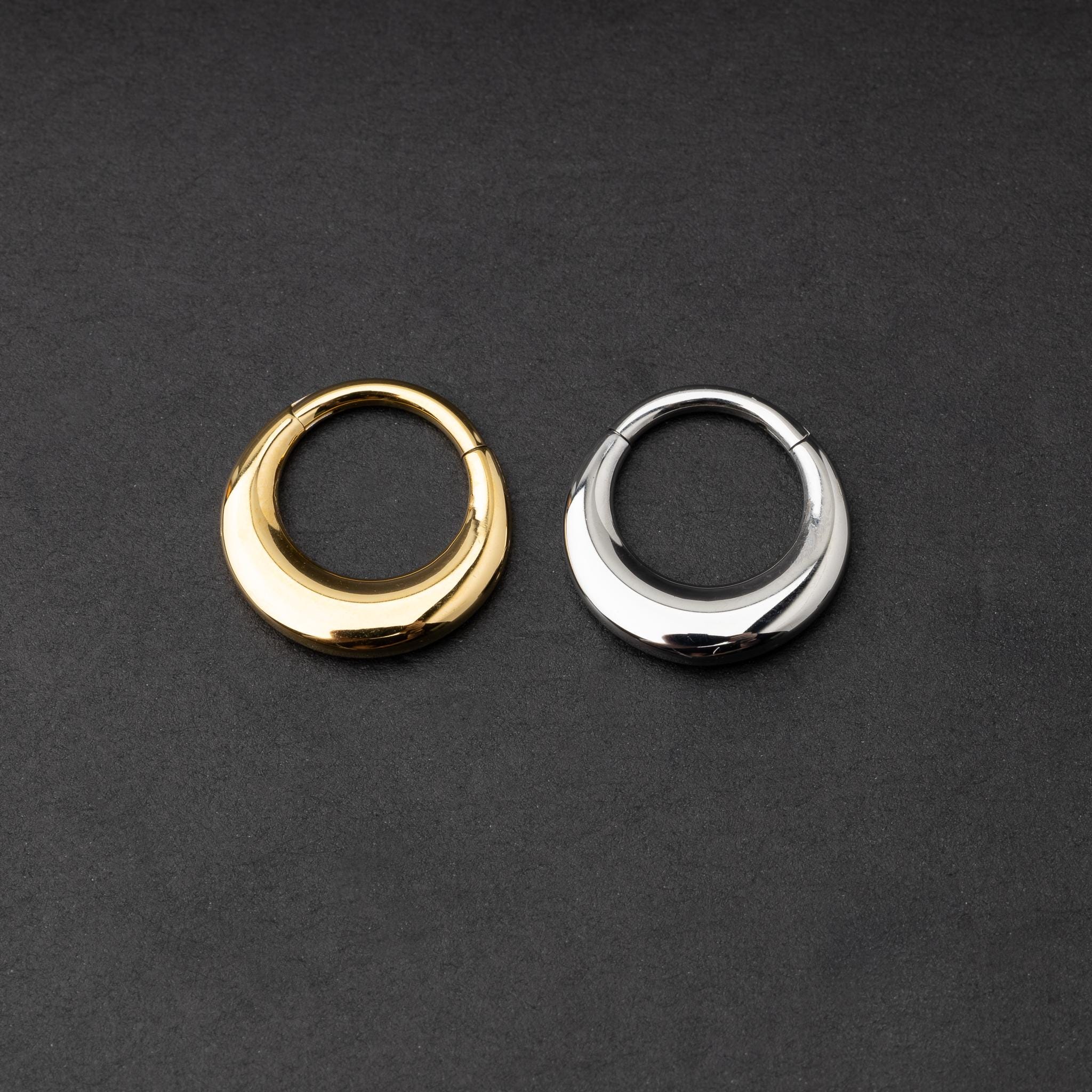 Crescent-shaped septum clickers, one in gold and one in silver, with a polished finish, displayed on a dark background.