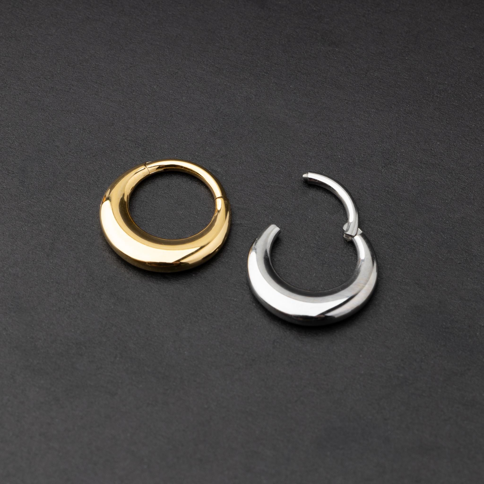Crescent-shaped septum clickers, one in gold and one in silver, with a polished finish, displayed on a dark background.