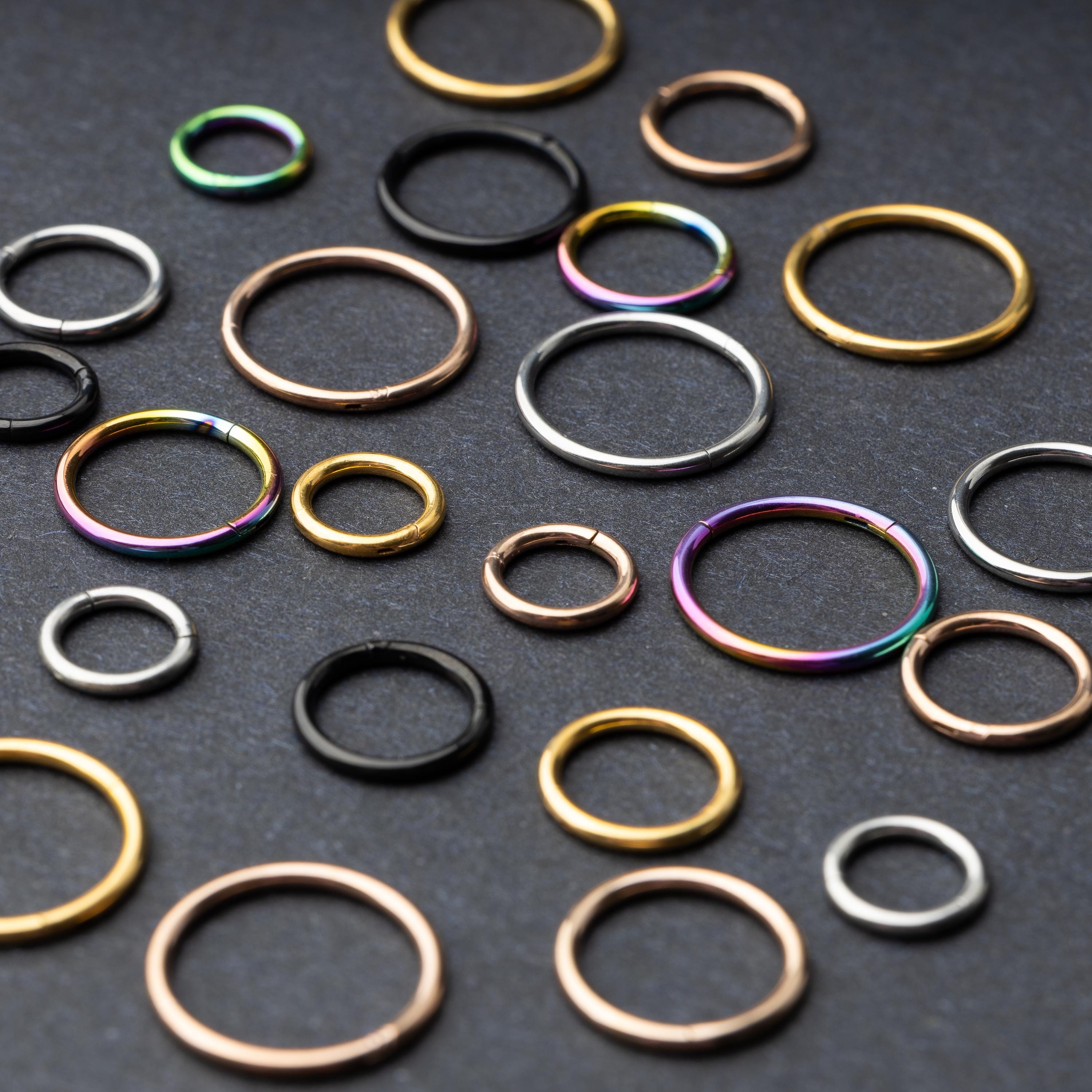 A variety of individually sold seamless clicker hoops in different sizes and finishes, including gold, rose gold, silver, black, and rainbow, displayed on a dark background.