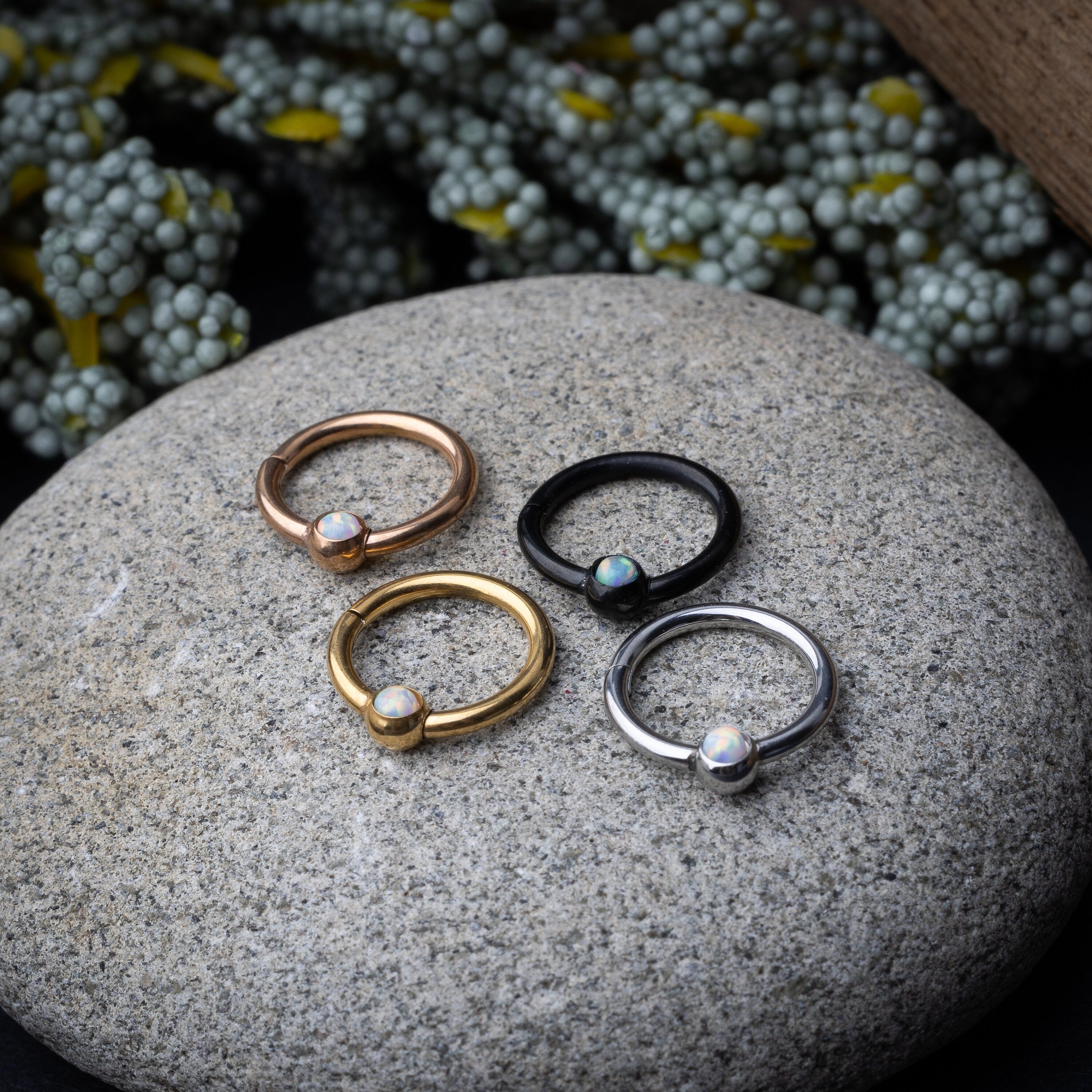Four seamless rings in black, gold, rose gold, and silver finishes, each featuring an opalescent bead, displayed on a stone background.