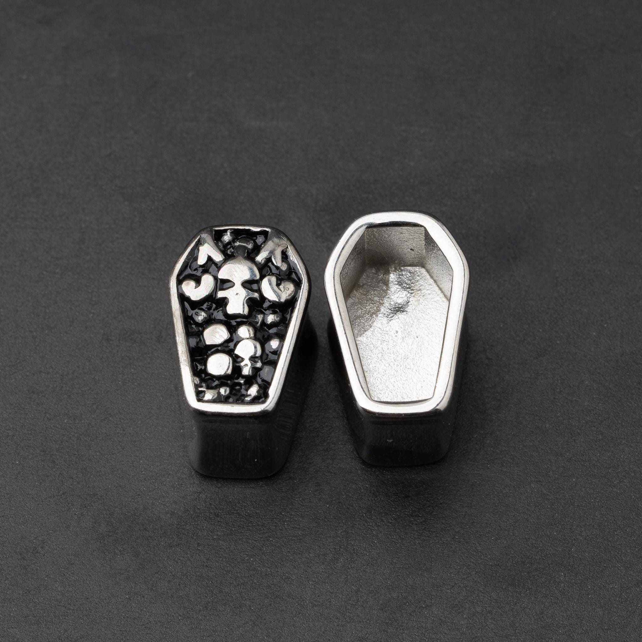 A pair of coffin-shaped stainless steel ear plugs with intricate skeleton designs, displayed on a dark background.