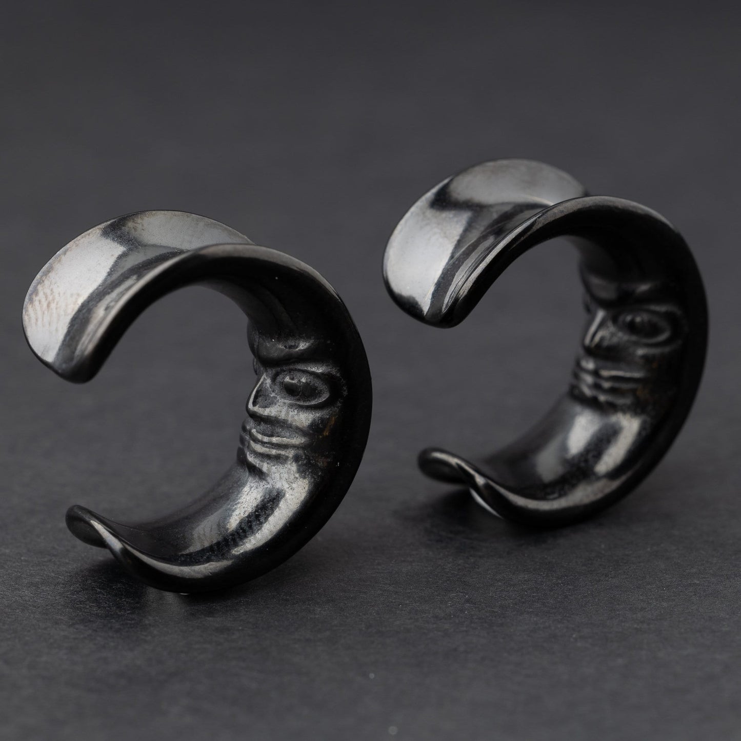 Close-up of a pair of black crescent moon-shaped ear weights with a detailed face design, displayed on a dark background.