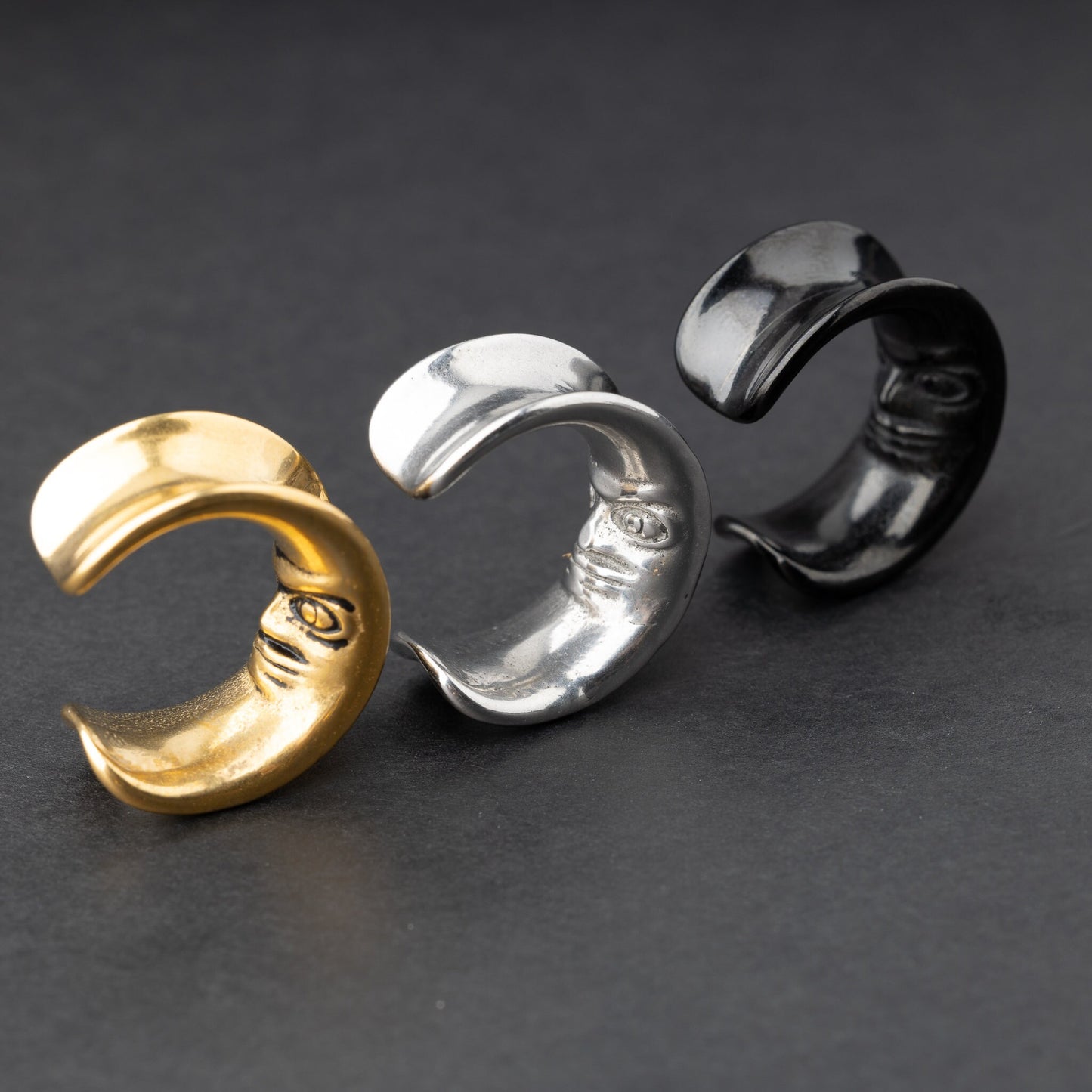 Close-up of a gold, black and silver crescent moon-shaped ear weights with a detailed face design, displayed on a dark background.