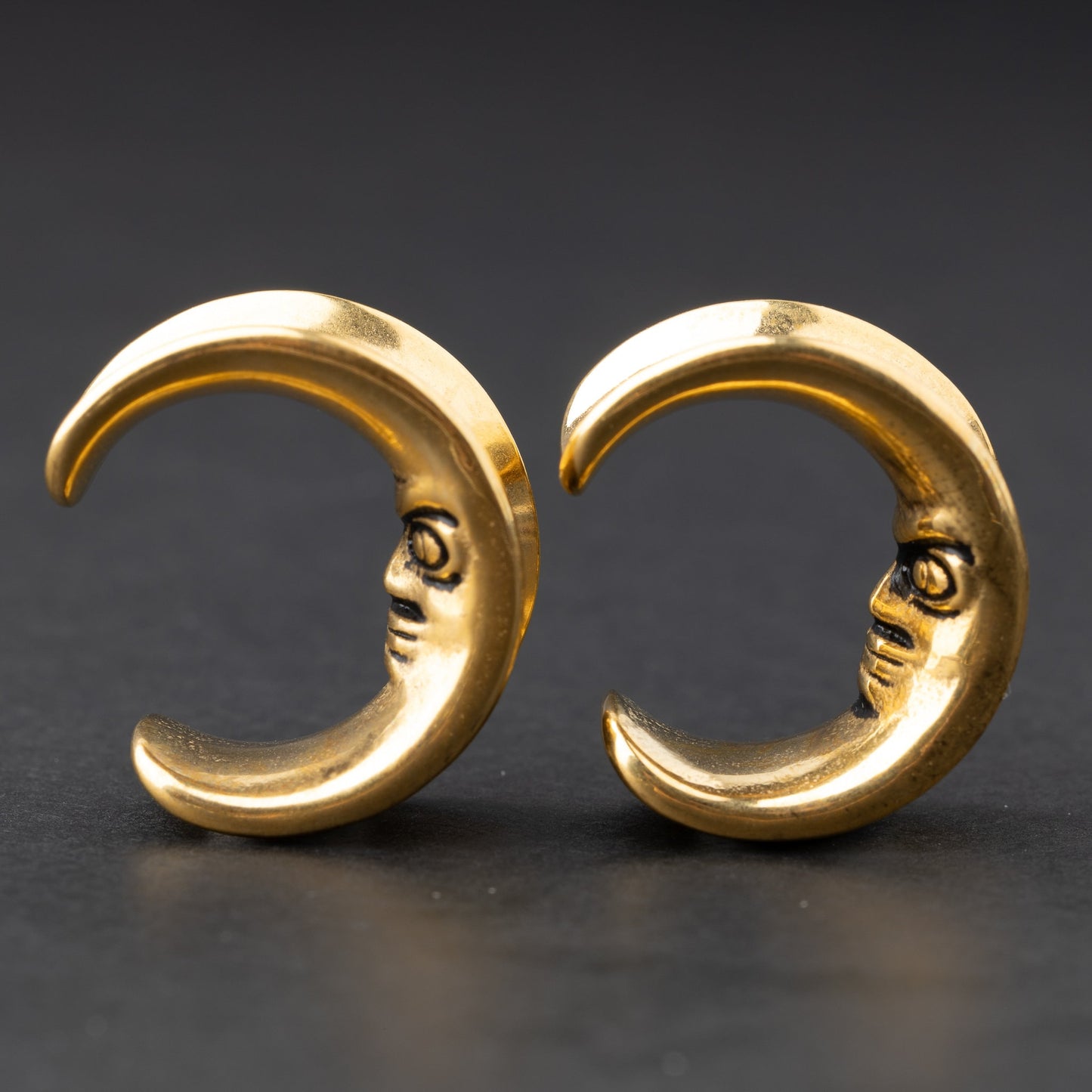 Close-up of a pair of gold crescent moon-shaped ear weights with a detailed face design, displayed on a dark background.