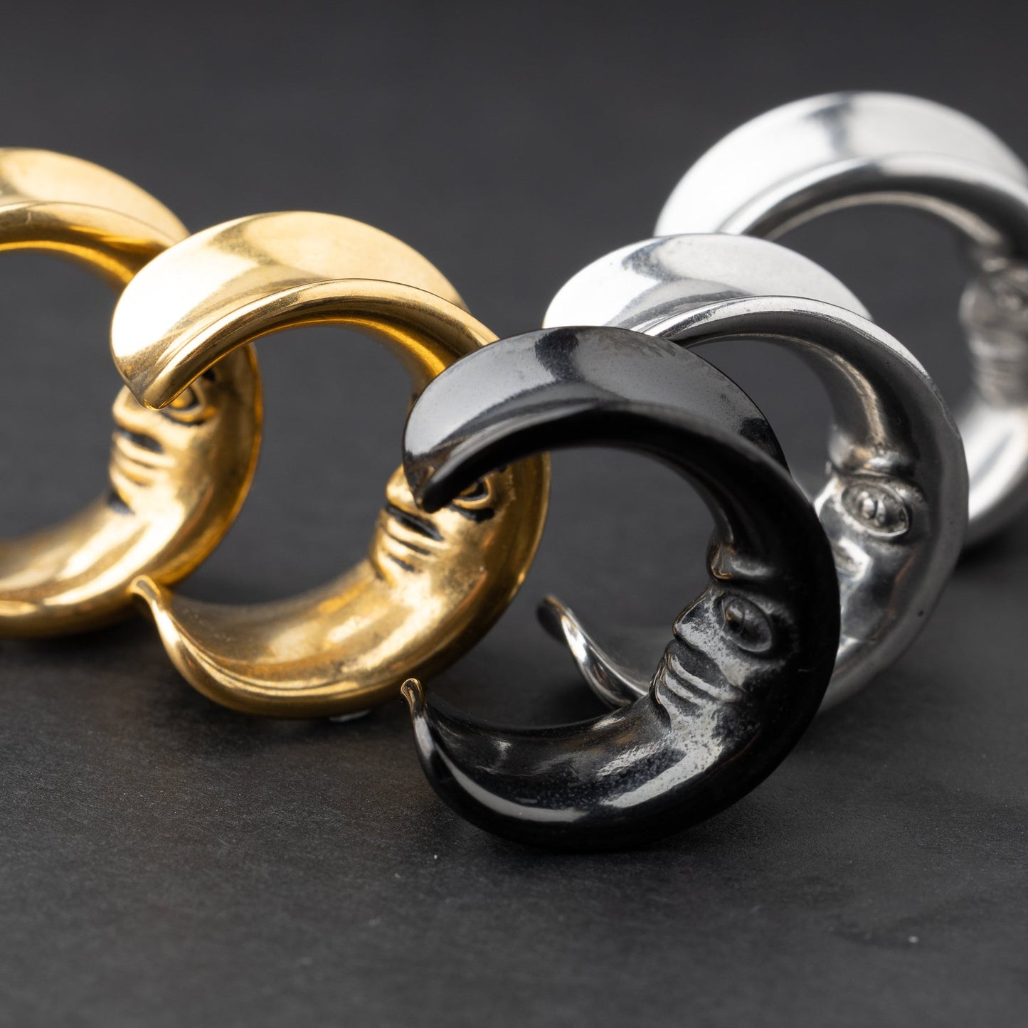 Close-up of a gold, black and silver crescent moon-shaped ear weights with a detailed face design, displayed on a dark background.