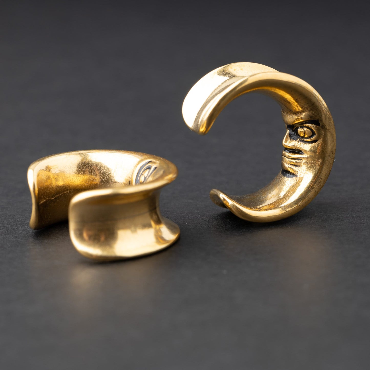Close-up of a pair of gold crescent moon-shaped ear weights with a detailed face design, displayed on a dark background.