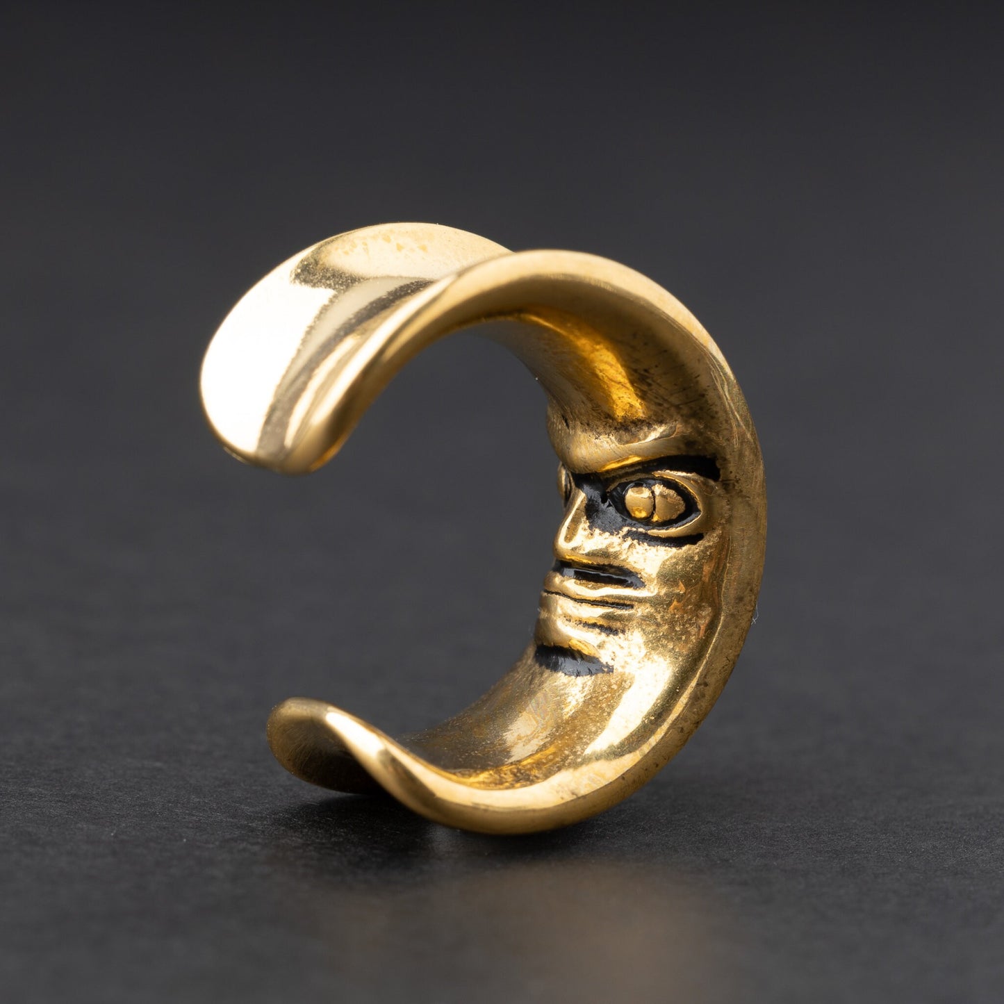 Close-up of a gold crescent moon-shaped ear weight with a detailed face design, displayed on a dark background.