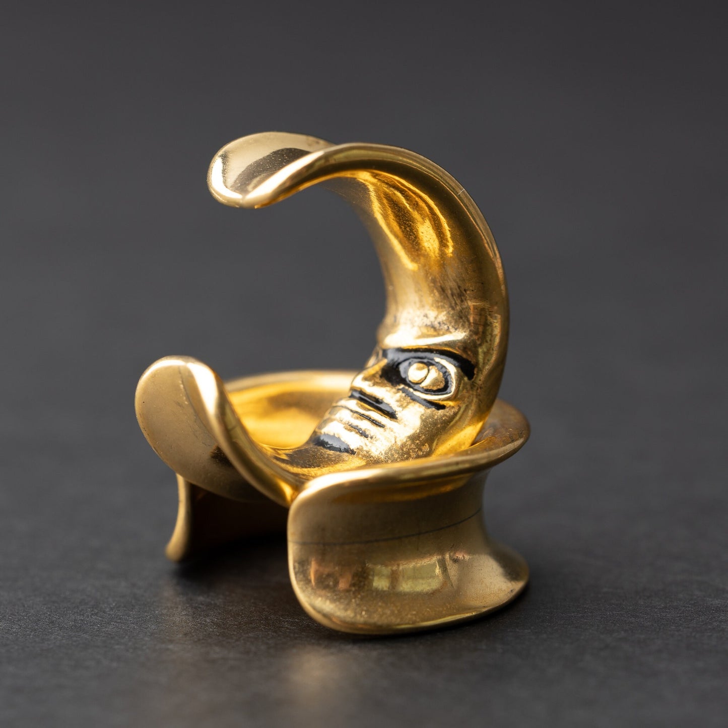 Close-up of a pair of gold crescent moon-shaped ear weights with a detailed face design, displayed on a dark background.