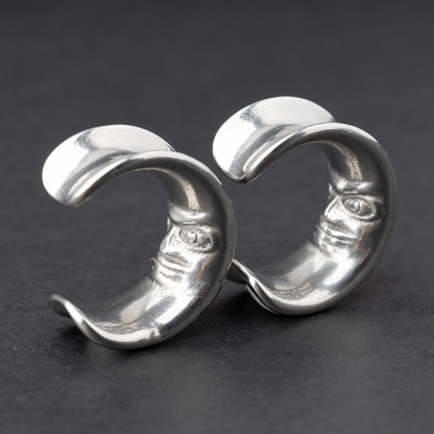 Close-up of a pair of silver crescent moon-shaped ear weights with a detailed face design, displayed on a dark background.