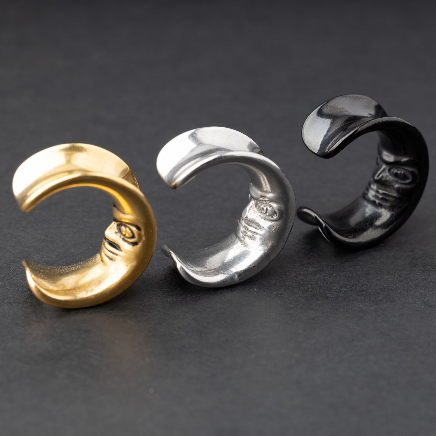 Close-up of a gold, black and silver crescent moon-shaped ear weights with a detailed face design, displayed on a dark background.