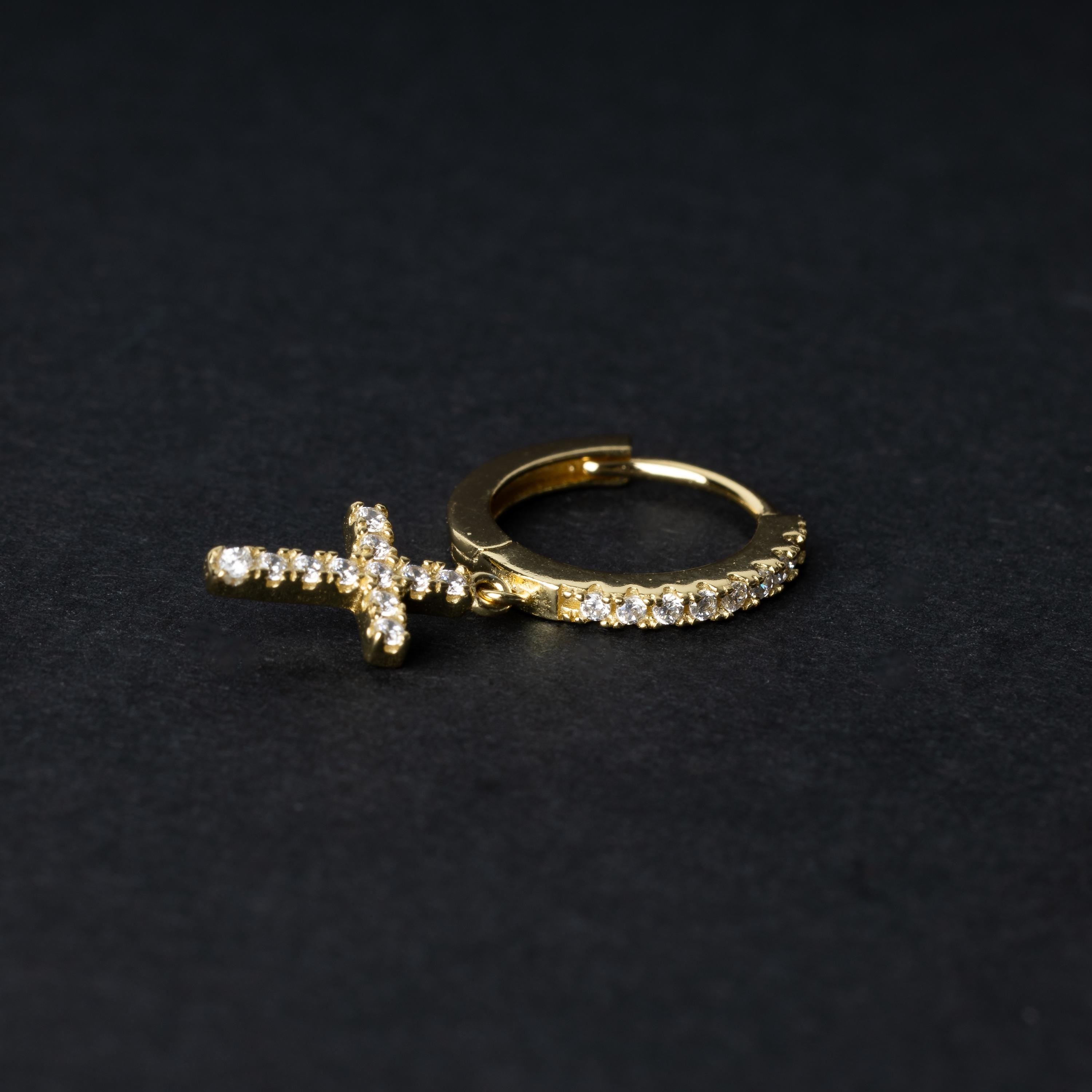 Gold hoop earring with cubic zirconia accents and a dangling cross charm, displayed on a dark background.