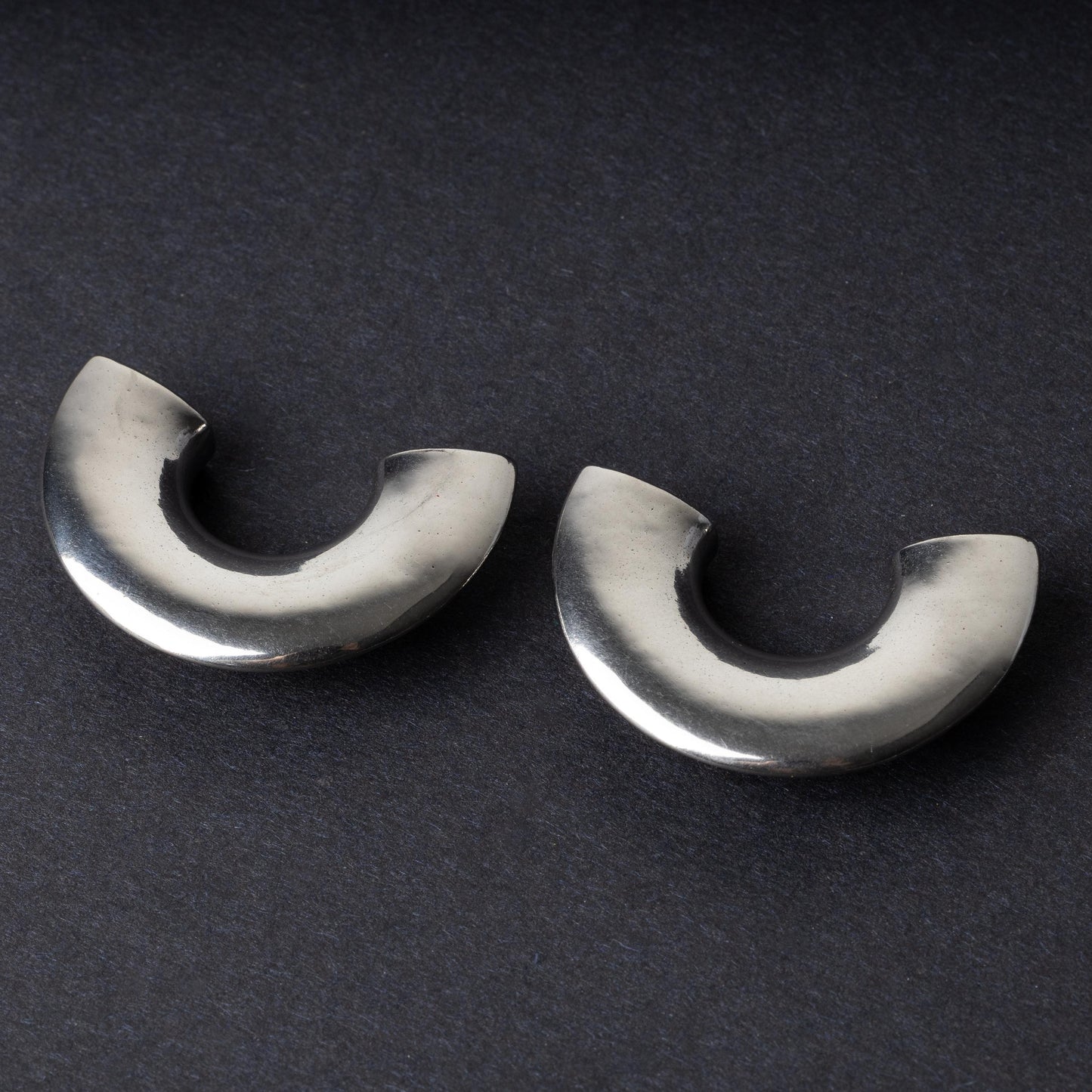 Close-up of two crescent-shaped stainless steel ear weights with a smooth, polished finish, displayed on a dark background.