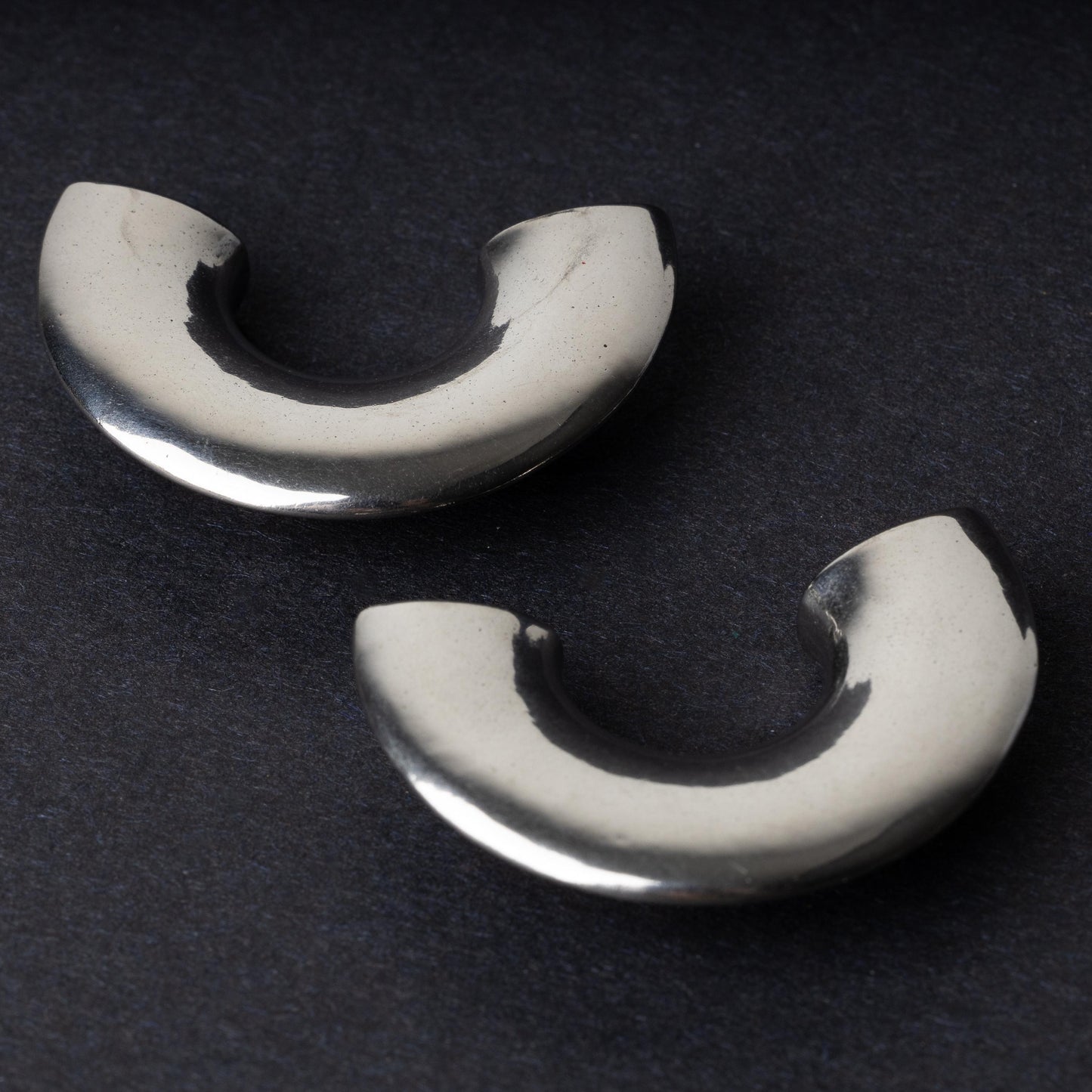 Close-up of two crescent-shaped stainless steel ear weights with a smooth, polished finish, displayed on a dark background.