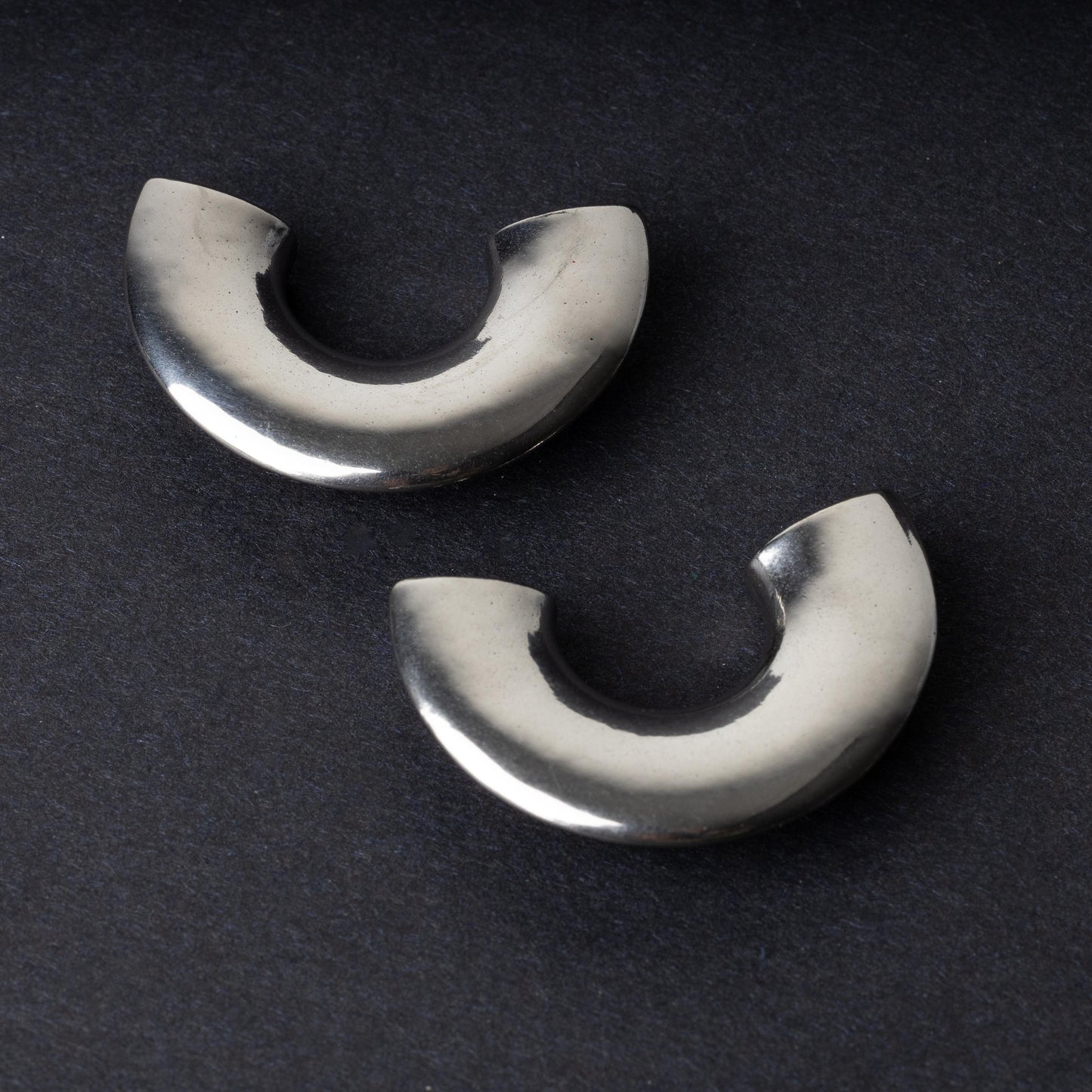 Close-up of two crescent-shaped stainless steel ear weights with a smooth, polished finish, displayed on a dark background.