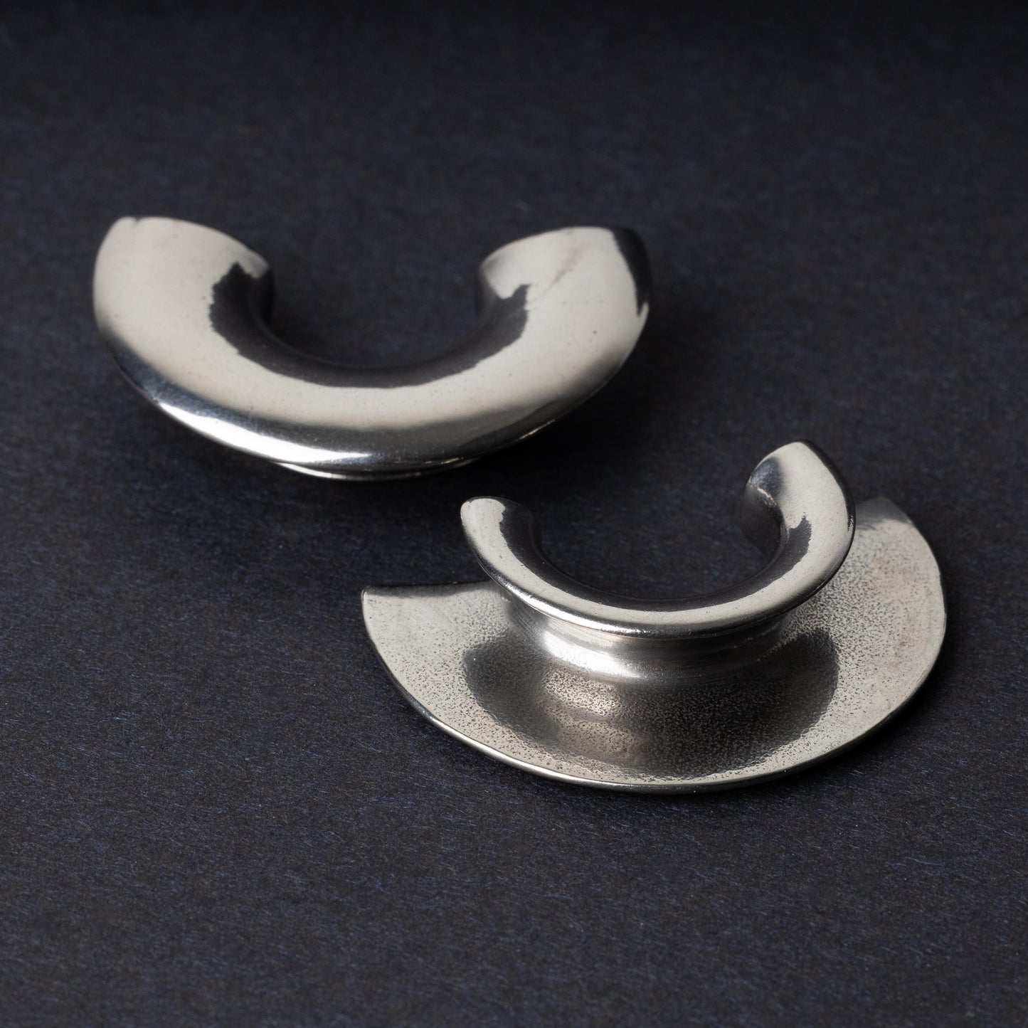 Close-up of two crescent-shaped stainless steel ear weights with a smooth, polished finish, displayed on a dark background.