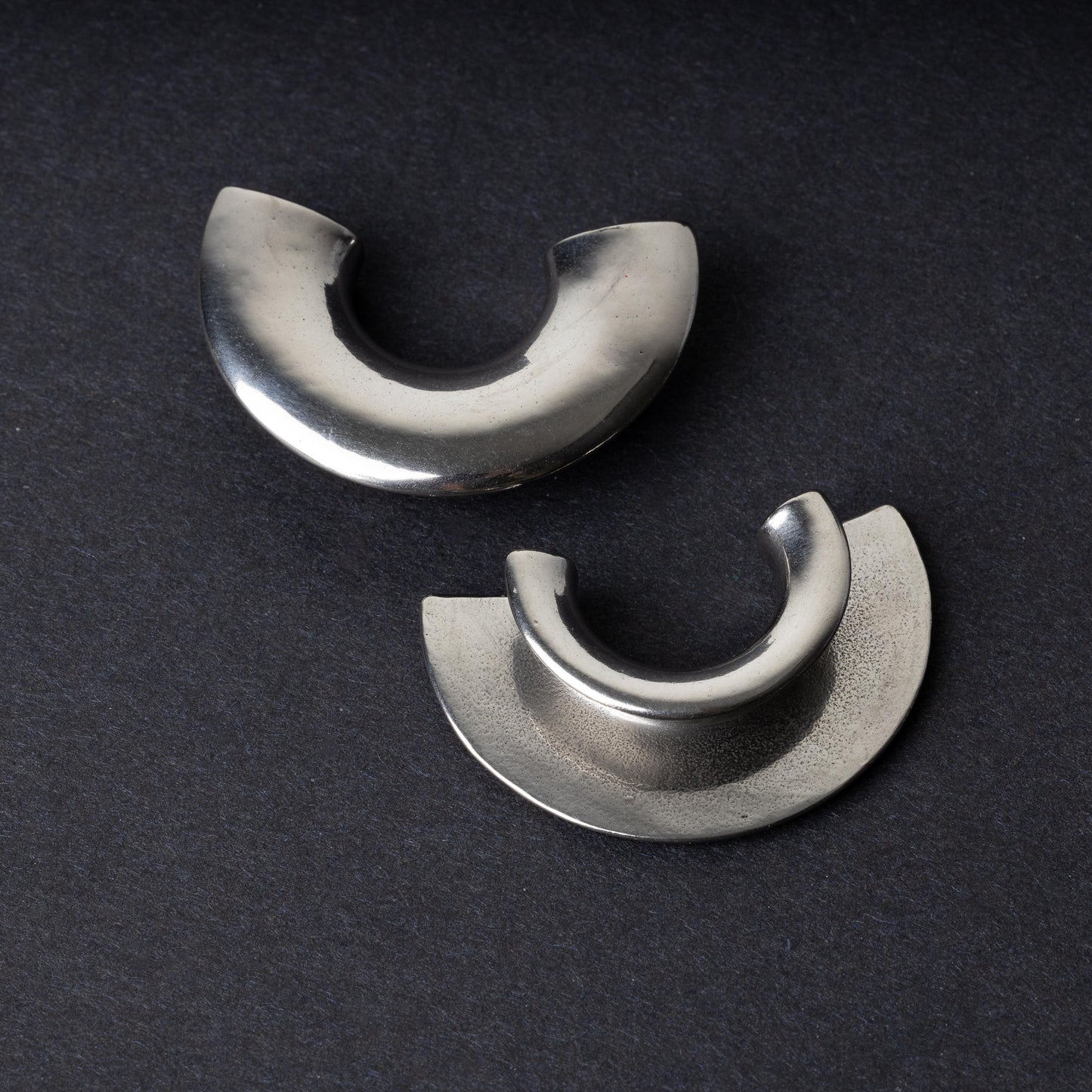 Close-up of two crescent-shaped stainless steel ear weights with a smooth, polished finish, displayed on a dark background.