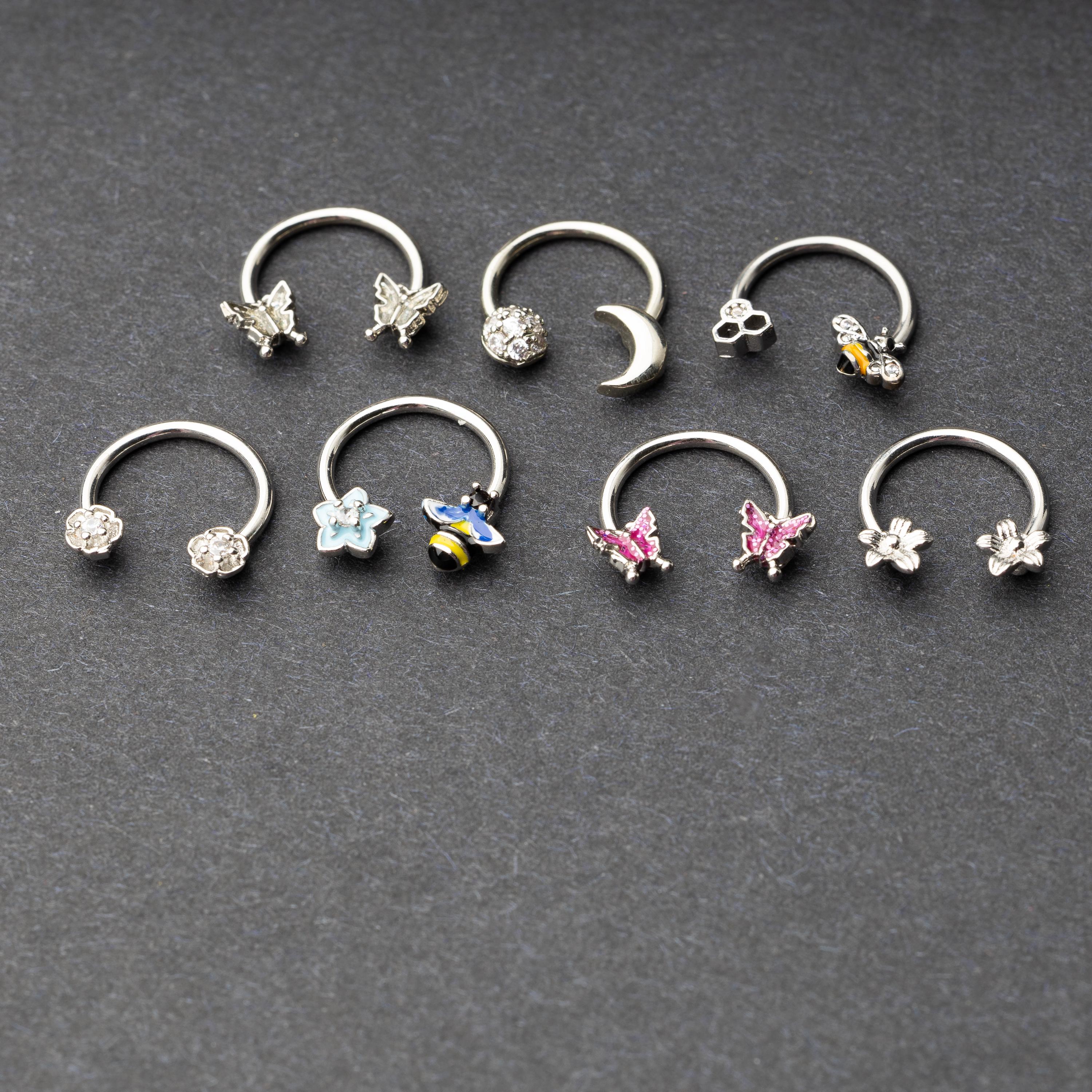 Silver horseshoe rings featuring various charms, including butterflies, bees, moons, and flowers, displayed on a dark background.