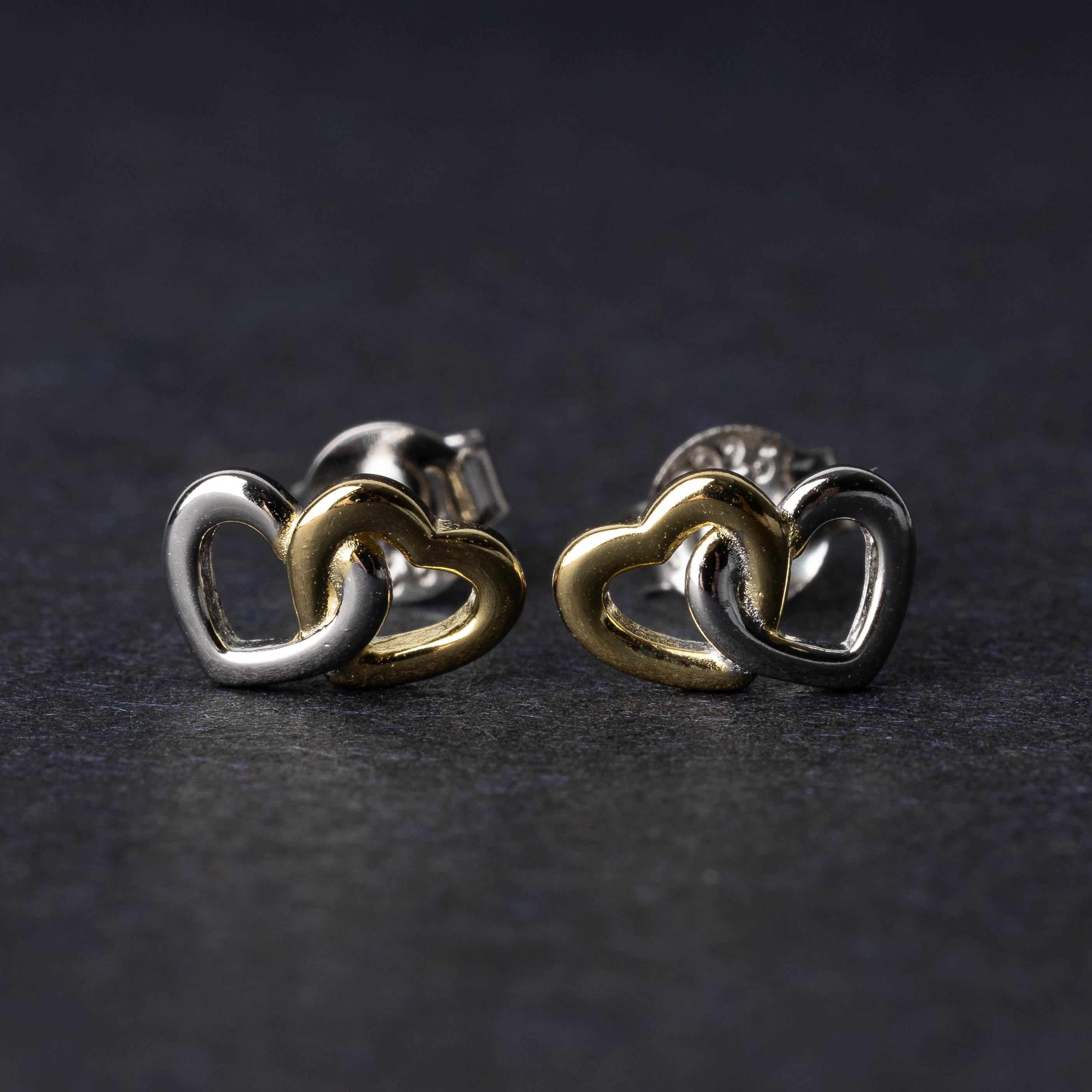 Close-up of interlocking silver and gold heart stud earrings with a polished finish, displayed on a dark background.