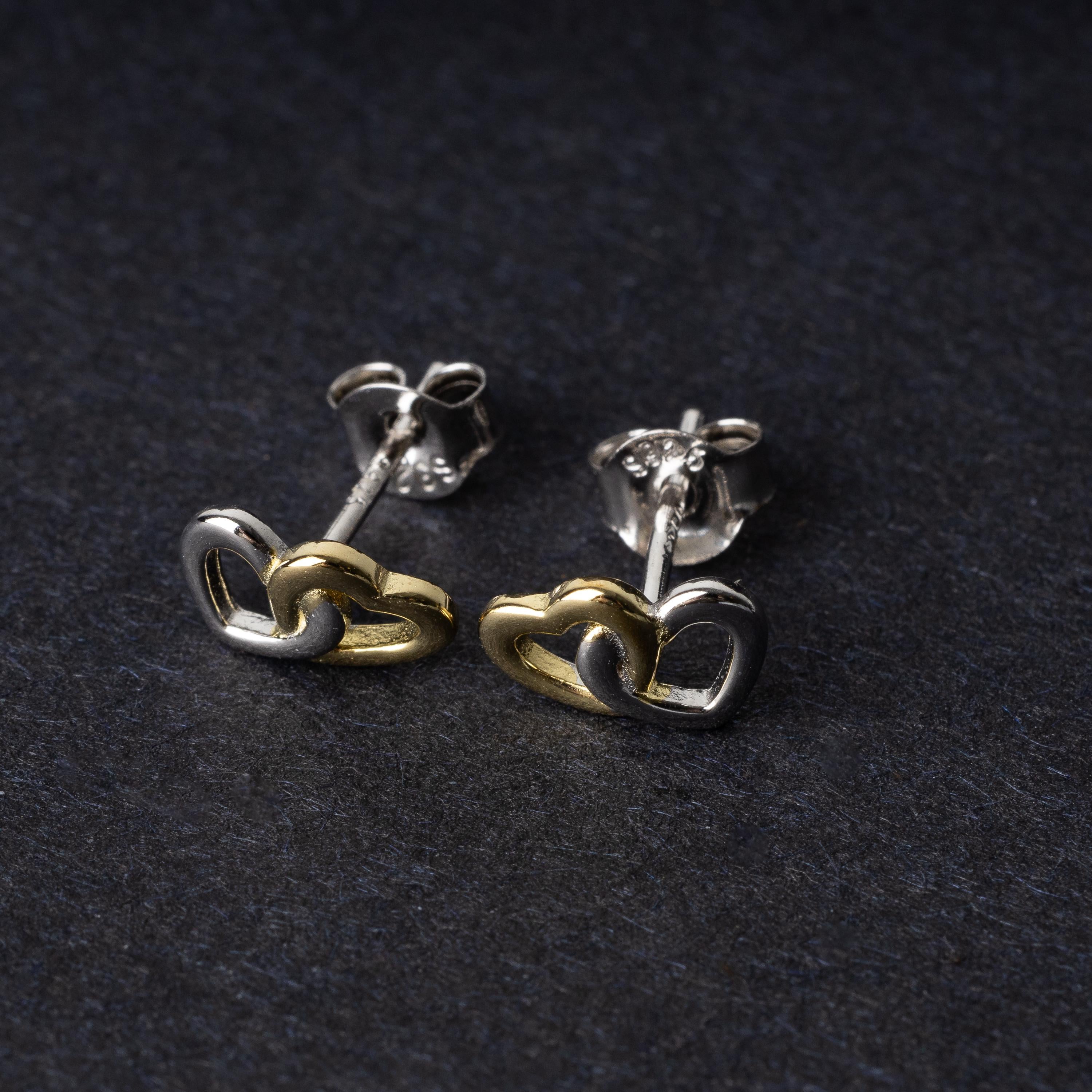 Close-up of interlocking silver and gold heart stud earrings with a polished finish, displayed on a dark background.