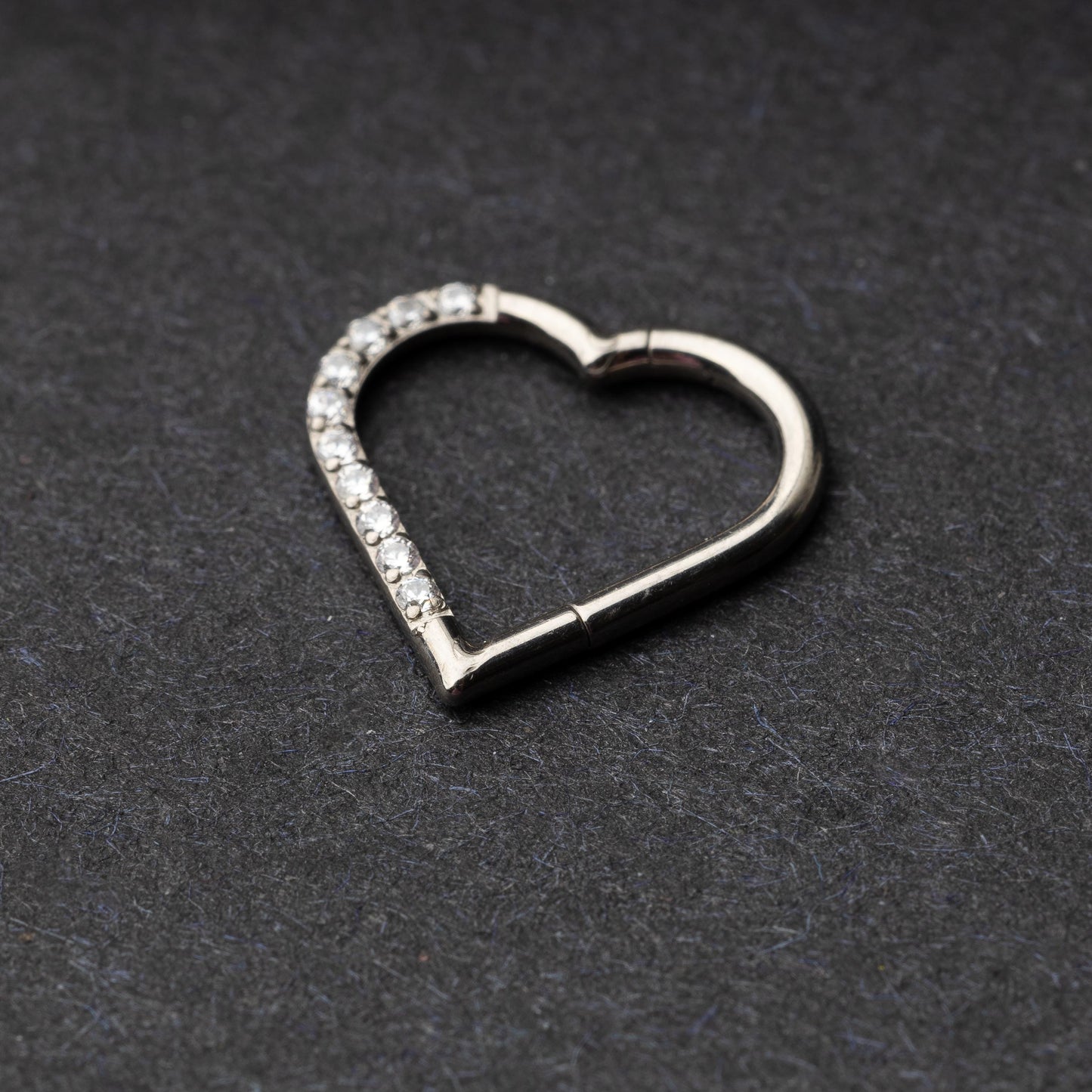 Heart-shaped daith clicker with crystal accents along one side, displayed on a dark textured background.
